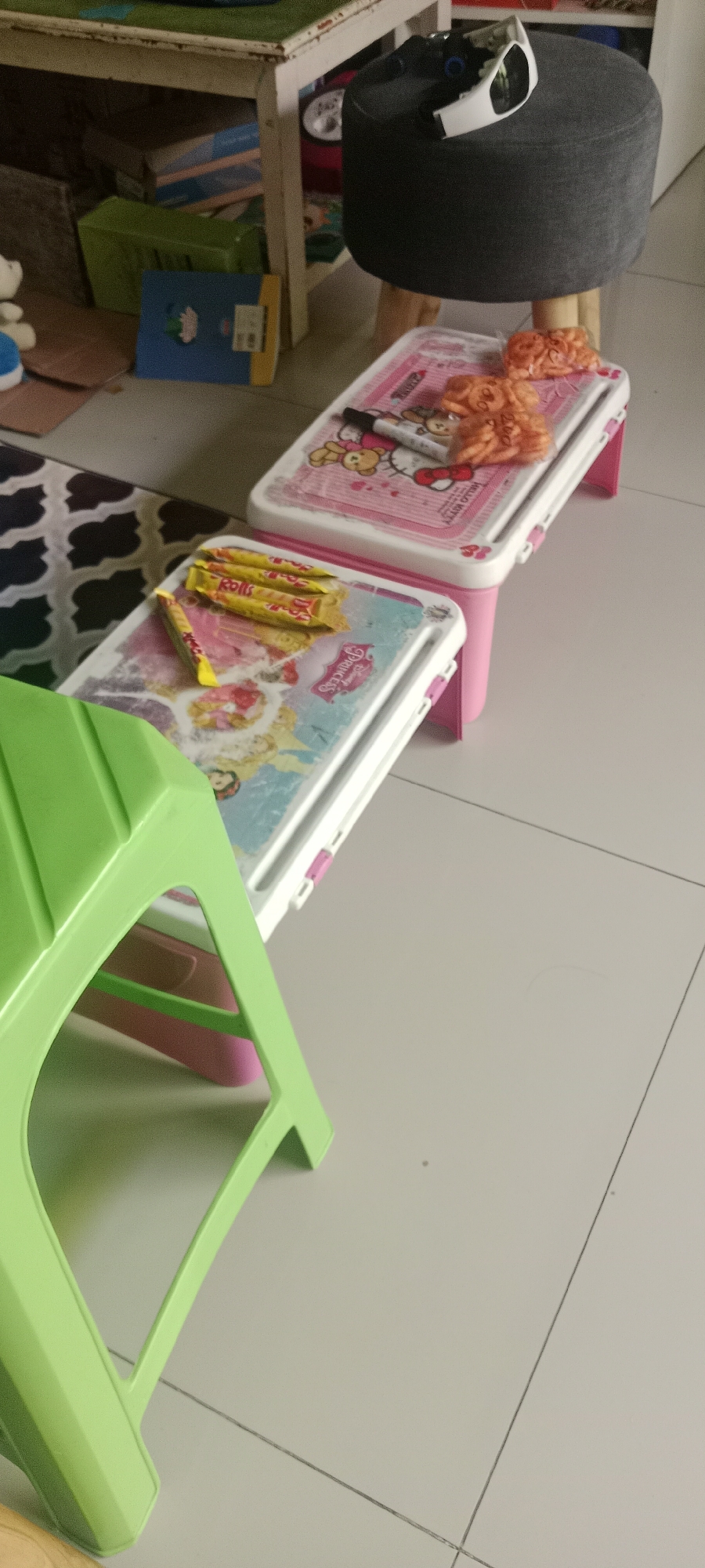 The image depicts a floor-level view of a room in Maguwoharjo, Indonesia. In the foreground, a bright green plastic stool is prominently displayed, its legs extending downwards. Behind the stool, two small, pink, foldable tables are arranged in a staggered fashion. The table closer to the foreground has several yellow-wrapped snack bars and a single yellow snack bar with red text on its surface, arranged on top. The table further back is adorned with a Hello Kitty design and features a marker pen and a small pile of orange, textured objects, possibly decorative items or snacks. In the background, partially visible, is a dark grey stool with wooden legs, topped with what appears to be a white and black electronic device, possibly a smartwatch or a remote control. Various storage boxes and children's books are stacked and placed on shelves and the floor under a white table in the background. The flooring is tiled and appears to be light grey with dark grout lines. The overall scene suggests a domestic environment, possibly a child's play area or study space.
