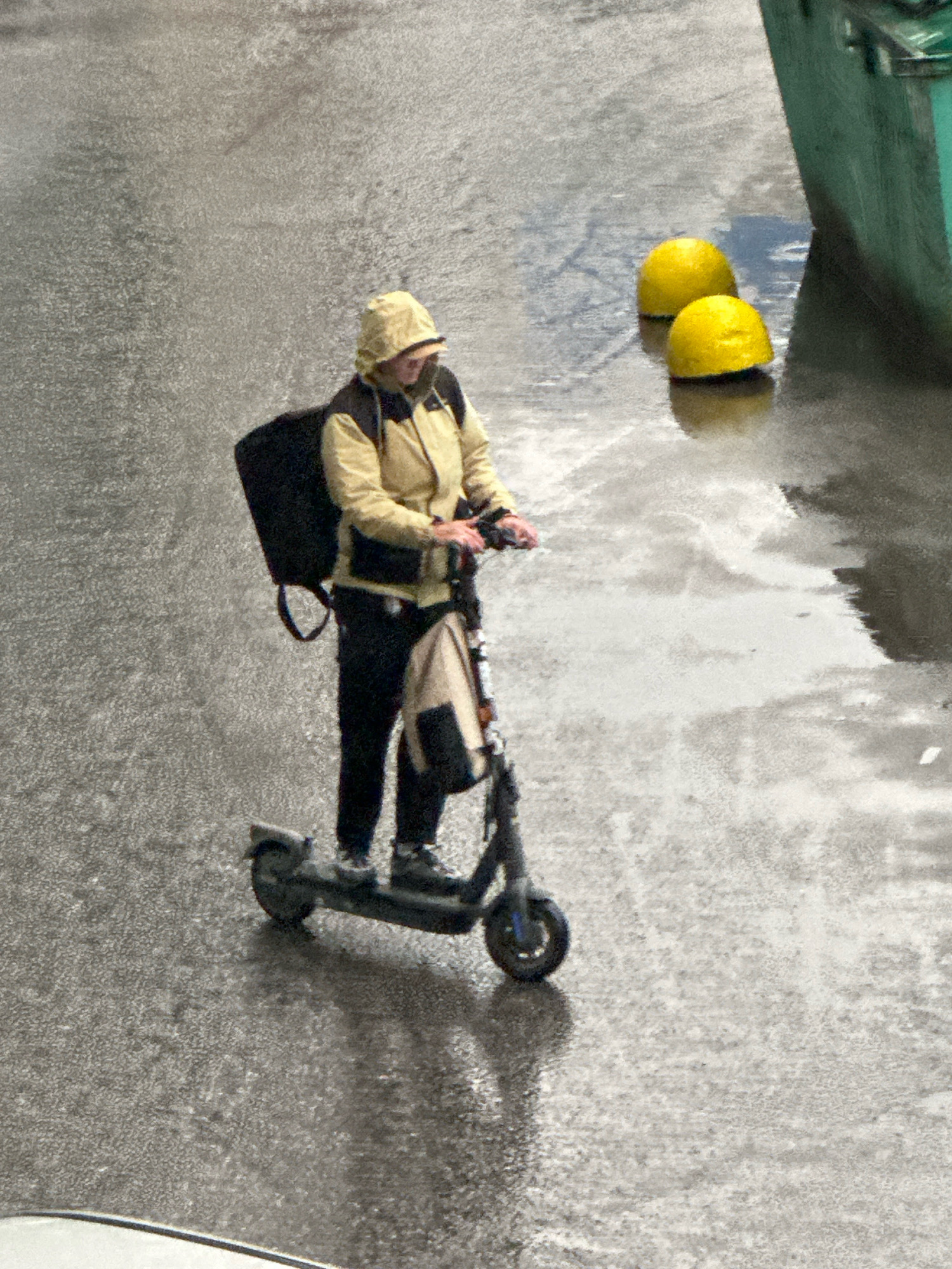 In Unknown, Russia, a person wearing a yellow hooded jacket, dark pants, and a face mask rides a dark-colored electric scooter on a wet, gray asphalt surface. The person has a large black bag strapped to their back and a smaller bag hanging from their shoulder. Two bright yellow buoys are partially submerged in water to the right of the scooter. A portion of a dark green structure is visible in the upper right corner of the image. The ground appears to be reflecting the light, indicating it is wet.