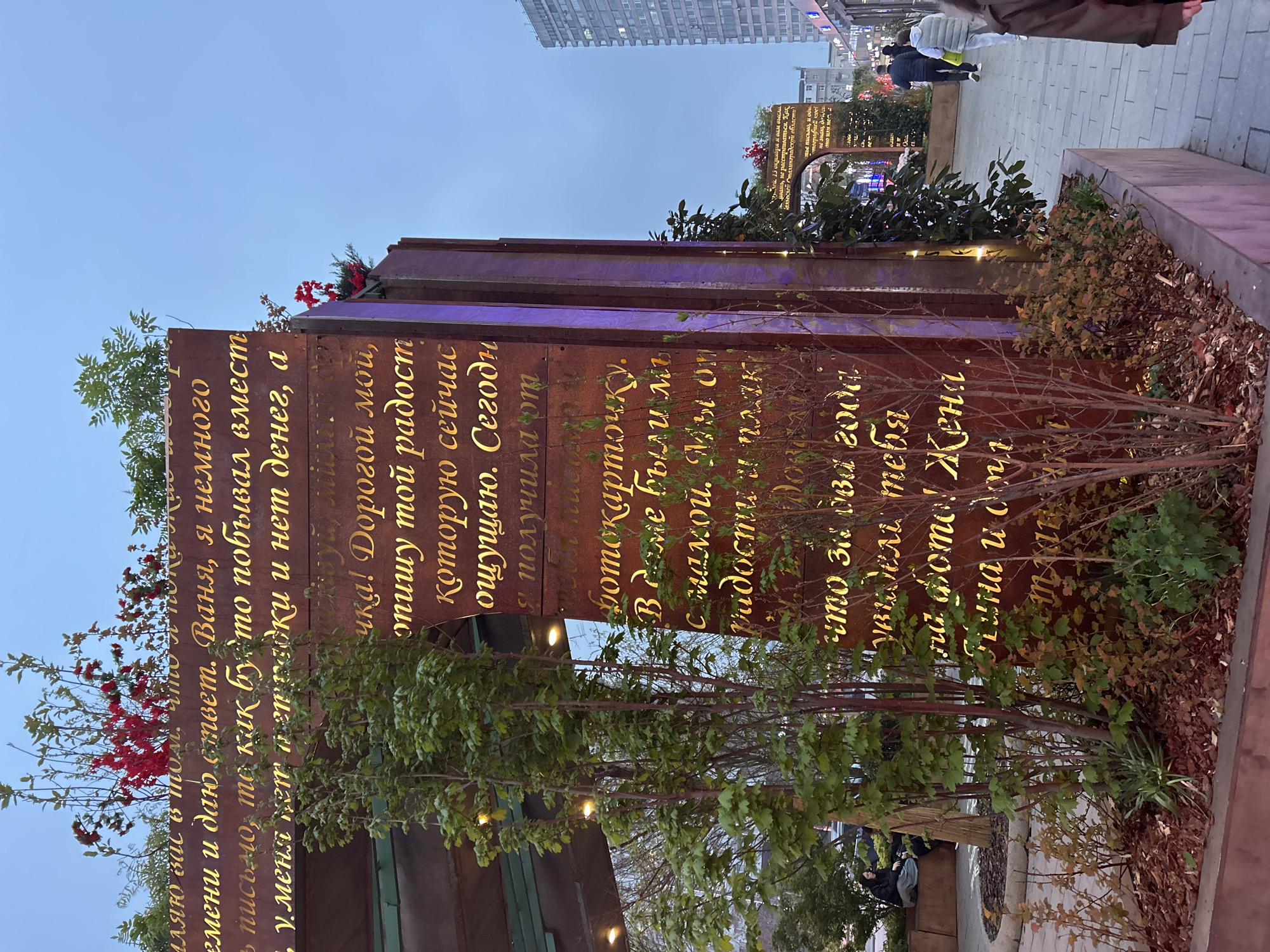 Here's a description of the image:
The image shows a weathered metal structure in an urban setting, likely a public artwork or memorial. The structure is composed of vertical metal slabs, with text in Russian etched onto the surfaces in a gold-colored font. The text appears to be a personal message or dedication.
The structure is partially surrounded by greenery, including small trees and bushes. This gives it a somewhat integrated appearance with the surrounding environment.
In the background, the blurred forms of people walking on a sidewalk and modern buildings are visible, indicating a cityscape. The sky is overcast, suggesting it could be dusk or a cloudy day.
The location appears to be a pedestrianized area within the city of Unknown, Unknown, as indicated by the people and buildings in the background.