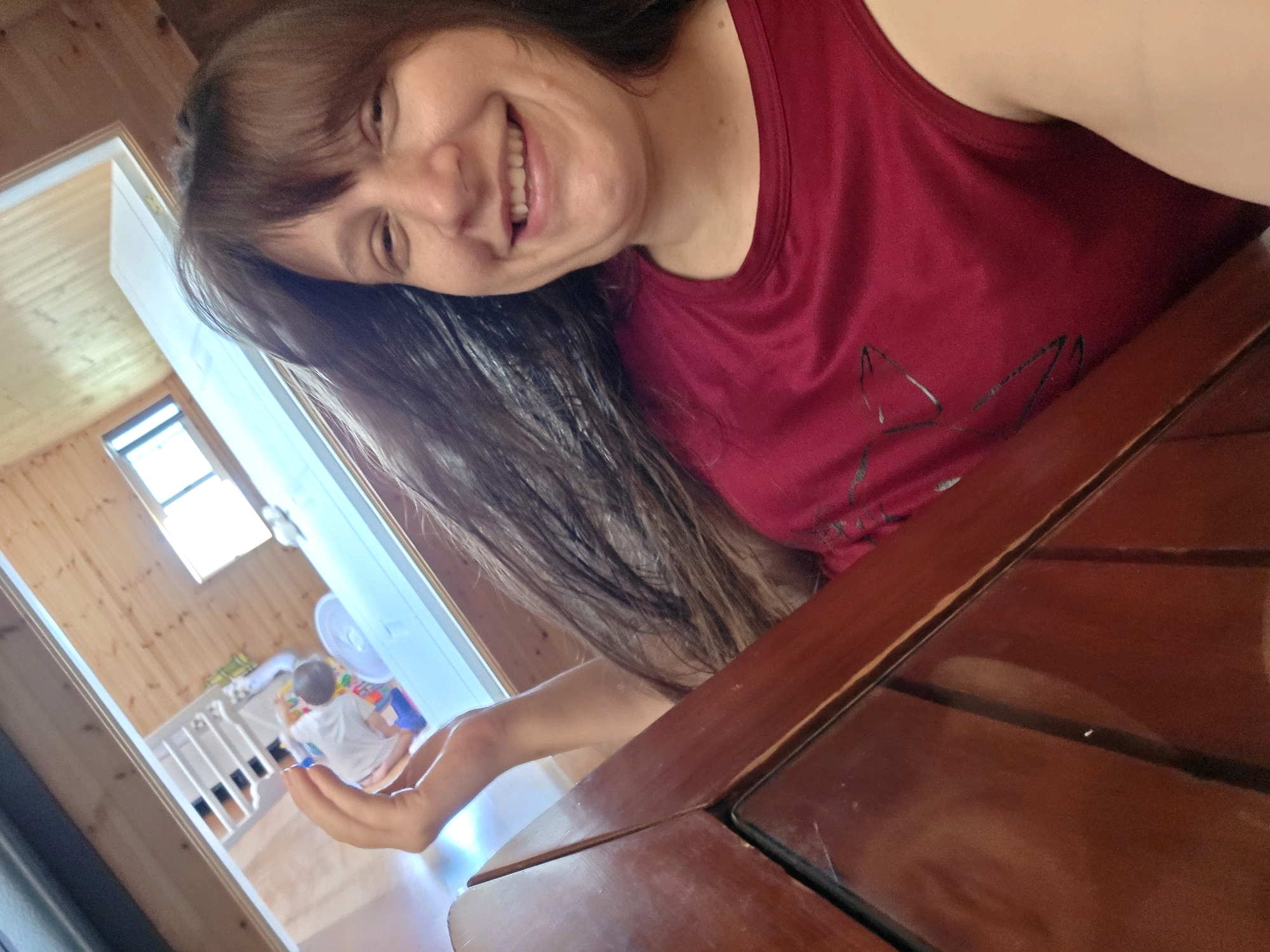 The image captures a close-up, selfie-style view of a woman with long, dark hair, wearing a red tank top. She is smiling and tilting her head. In the background, through an open doorway, a child is visible in a room with wooden walls. The child is seated and appears to be playing with toys. A white fan and a white crib-like structure are also present in the background. The foreground is dominated by a dark wooden table. The scene appears to be indoors, possibly a home.