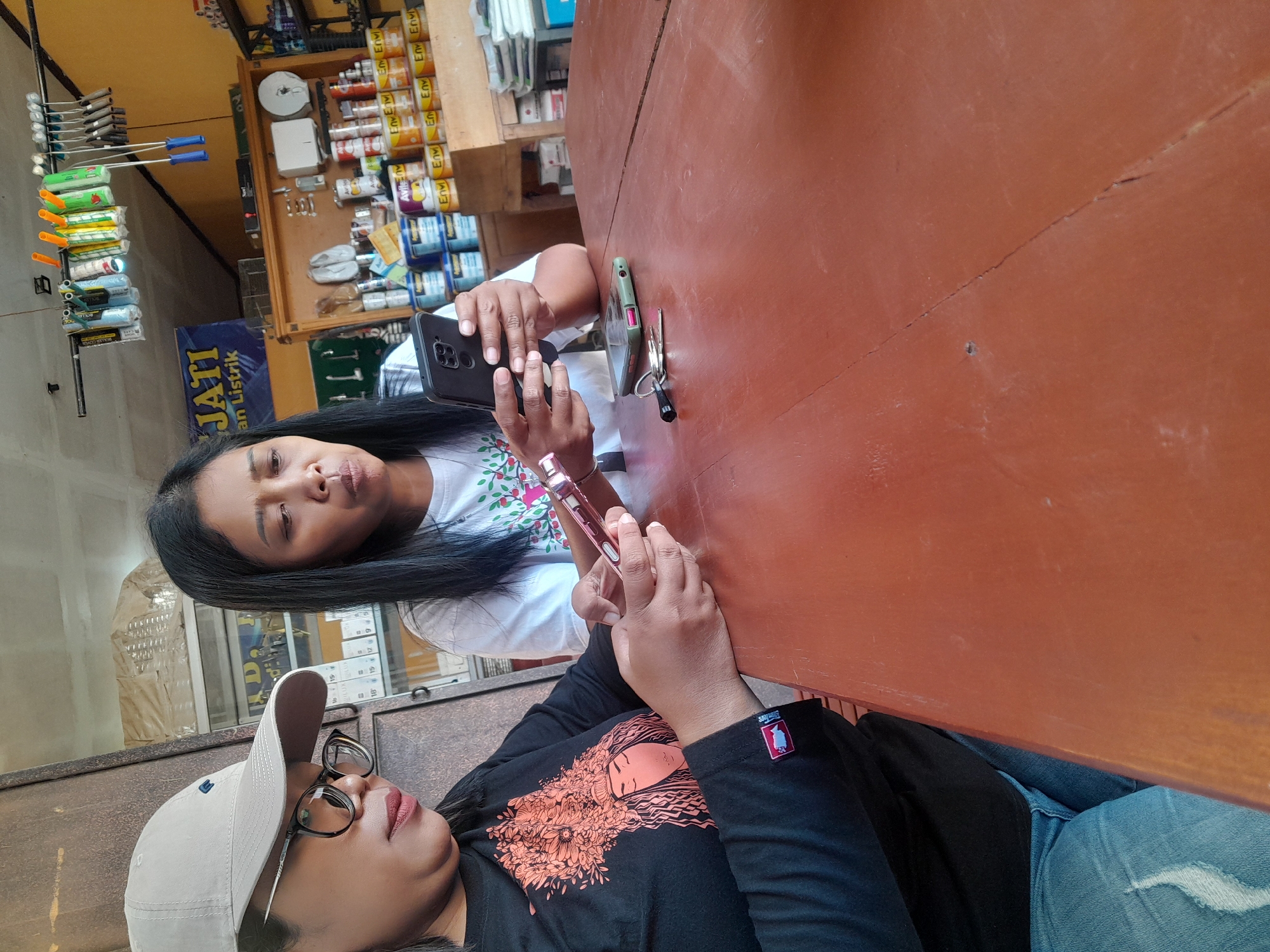 Two women are seated at a reddish-brown wooden table in what appears to be a shop in Purwoharjo, Indonesia. The woman on the left wears a black long-sleeved shirt with a graphic print and blue jeans, and a light-colored baseball cap. She is looking down at her phone, which she holds in her left hand. She wears glasses. The woman on the right wears a white t-shirt and has long black hair. She is looking at her phone, which she holds in her right hand. A second phone is lying on the table, face down. A set of keys is next to the second phone. In the background, shelves are stocked with cans of paint and other supplies. A sign with the words "JATI AN LISTRIK" is visible on the wall. The ceiling is visible, with some pipes and electrical fixtures.