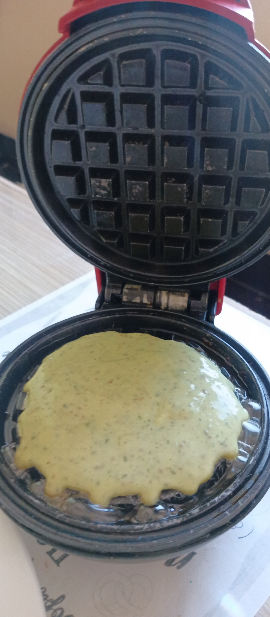An open, red and black waffle maker is shown from a high angle, with the top plate lifted. A circular blob of light green, speckled batter fills the center of the bottom waffle plate, just poured and spreading to the edges. The batter has small, dark specks throughout, suggesting ingredients like herbs or vegetables. The bottom plate appears to be well-greased, with some liquid visible around the batter. The top plate is clean, displaying its square grid pattern. The hinges connecting the top and bottom parts of the waffle maker are visible, showing some residue or wear. The background consists of a light-colored surface, possibly a countertop or placemat, with blurry, dark text and a pretzel-like design partially visible beneath the waffle maker. The image captures the moment before a waffle is cooked, with the batter ready to be pressed.
