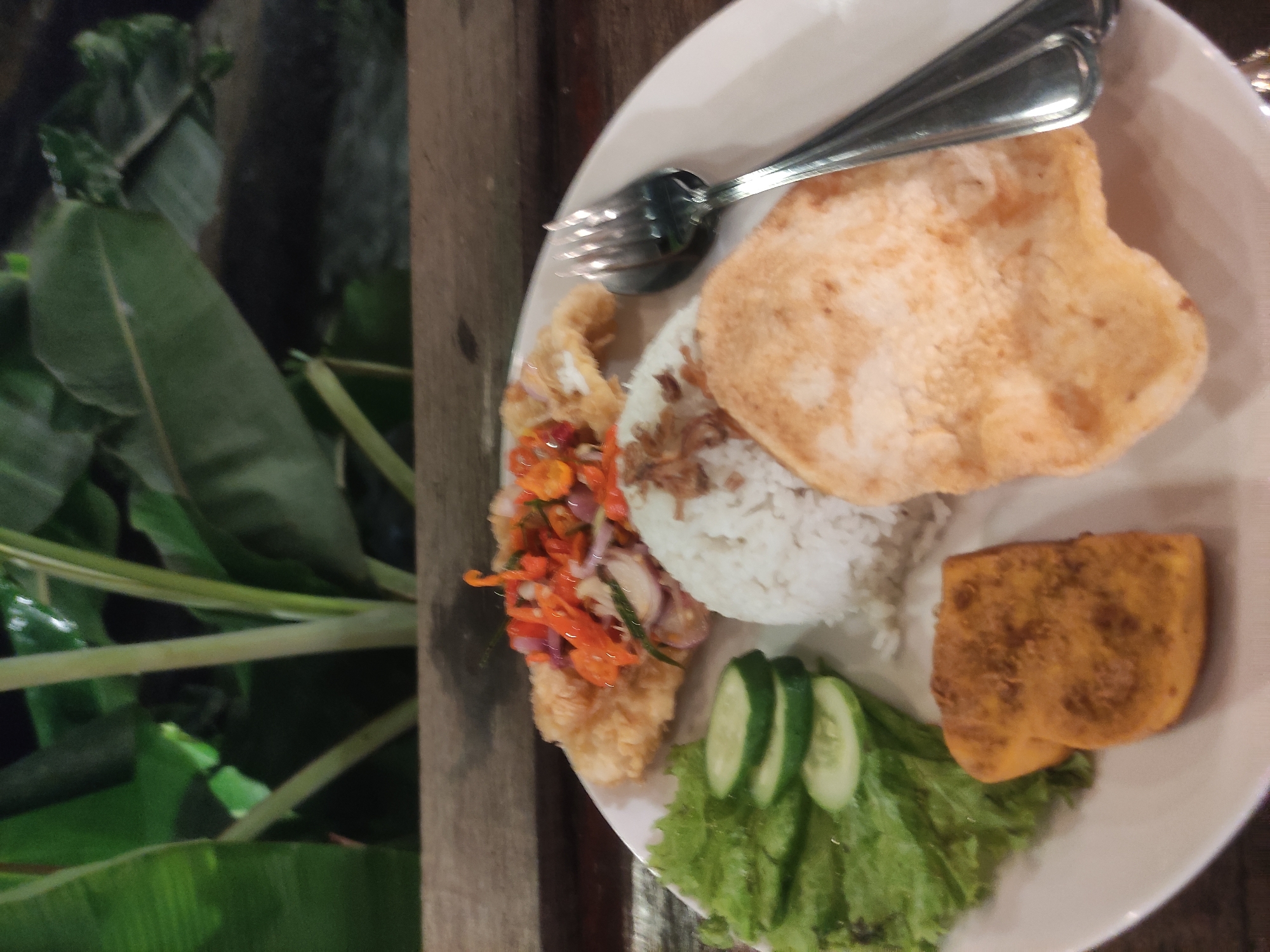 The image shows a plate of Indonesian food, likely photographed in or near Babakan Madang, Indonesia, given the prevalence of this cuisine in the region. The plate contains a serving of white rice, fried tofu, some pieces of cucumber and lettuce, a fried cracker, and what appears to be fried chicken topped with a red chili sauce. A spoon and fork are placed on the top right corner of the plate. The food is set on a wooden table or surface, and a green, leafy plant is visible to the left of the plate. The lighting is somewhat dim, suggesting an indoor or evening setting.