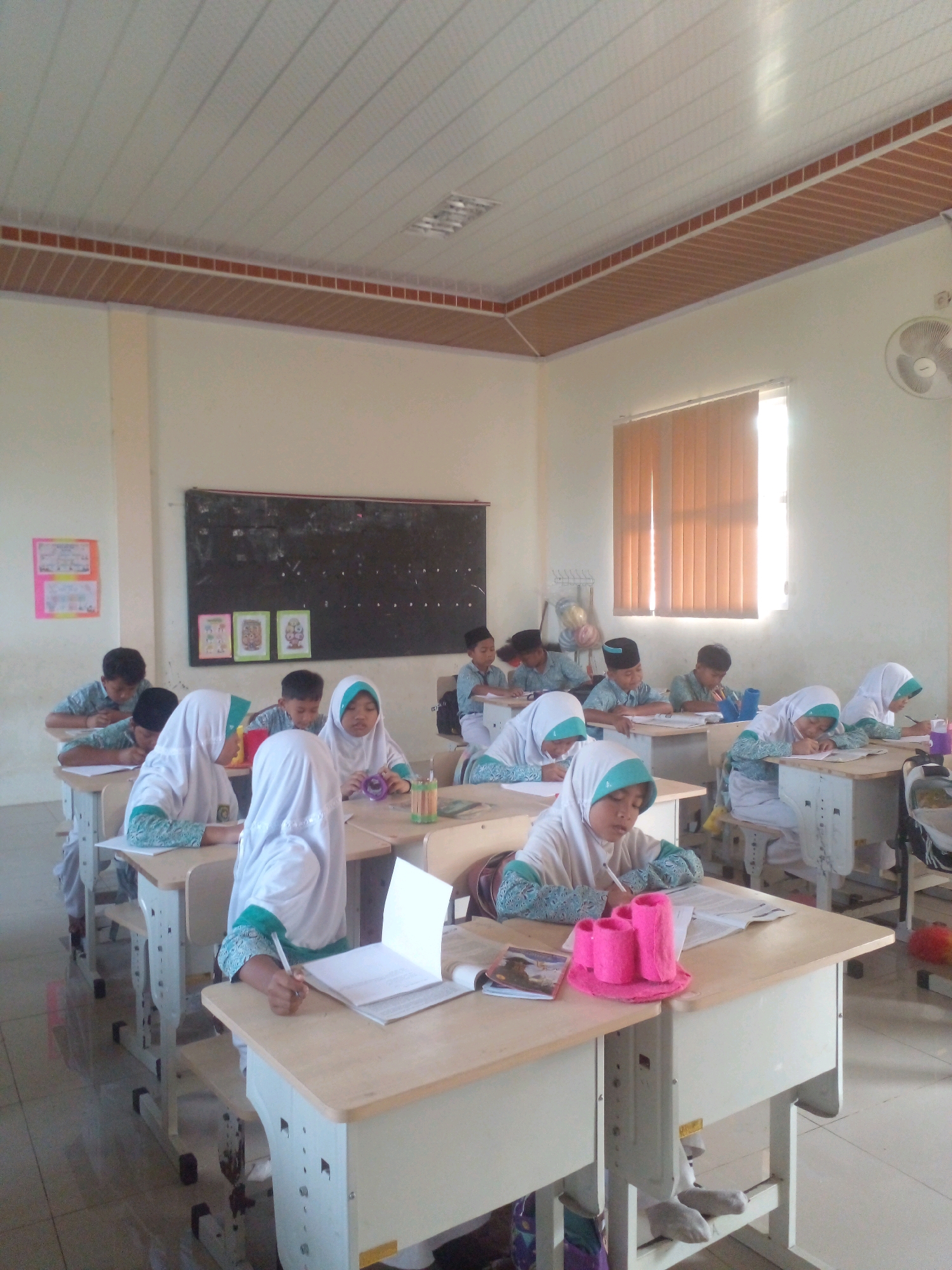 The image depicts a classroom scene in what is presumed to be an educational institution in Banyuputih, Indonesia. The room is brightly lit by natural light streaming from a large window covered by vertical blinds. The ceiling is paneled with white and wood-toned planks, with a fluorescent light fixture visible. Along the walls, there are white walls, a blackboard, and a colorful educational poster. The floor appears to be tiled.
Several young students, likely of elementary school age, are seated at individual wooden desks arranged in rows. The majority of the students are wearing white hijabs and matching long-sleeved tops with teal accents. The boys are wearing caps. The students are engaged in various activities, primarily writing or reading from notebooks and books on their desks. Some students are focused intently on their work, while others appear to be looking around the room.
On the desks, there are notebooks, pens, pencils, and some small containers. One desk in the foreground holds a pink stationary organizer. A fan is mounted on the wall in the background. The overall atmosphere suggests a quiet and focused learning environment.
