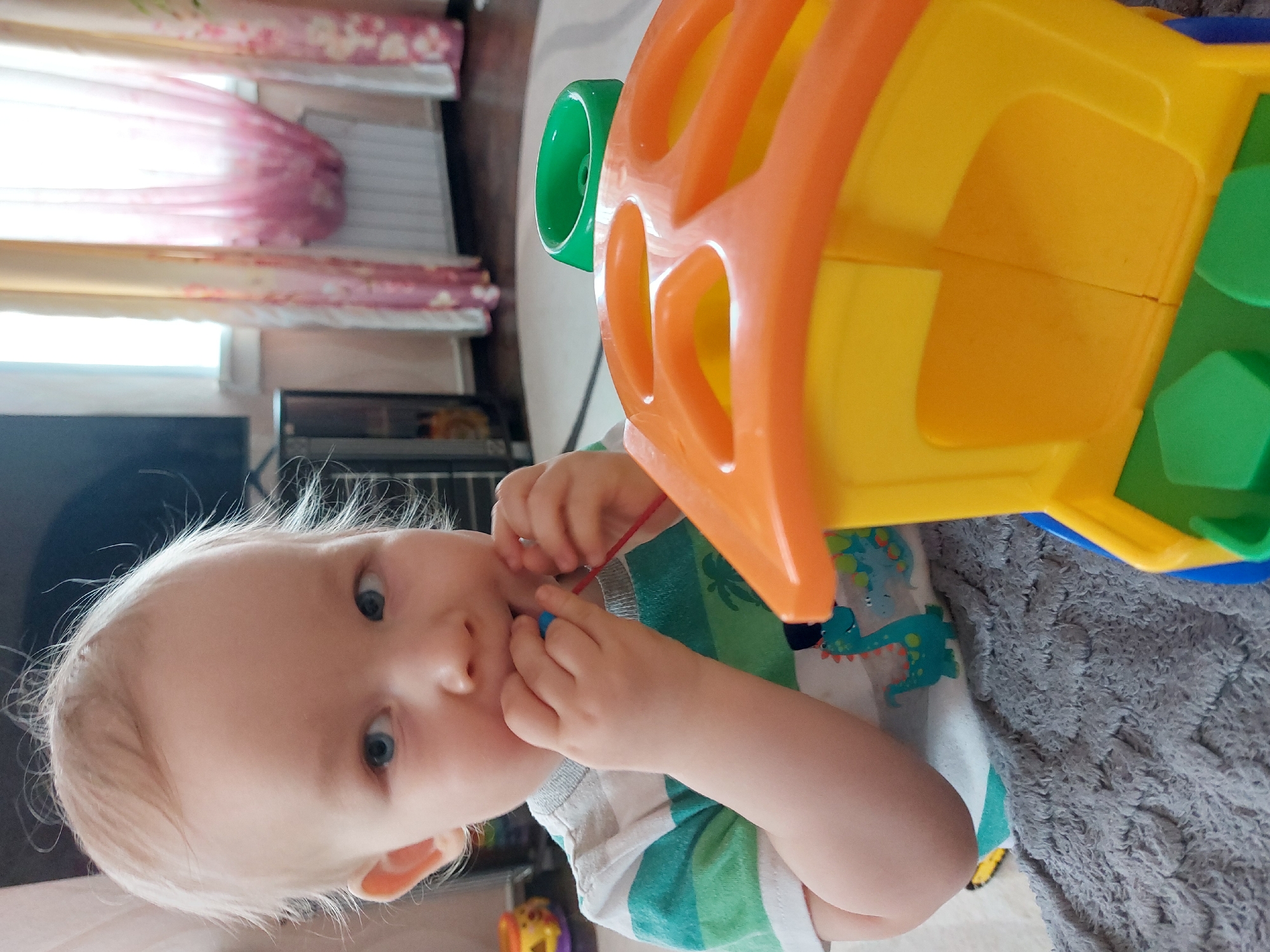 A young child with light hair and blue eyes is looking directly at the camera. The child is holding a blue toy in their hands, near their mouth. The child is wearing a striped green and white shirt with a pattern of blue dinosaurs on the chest. A large, colorful plastic toy with orange, yellow, and green components is positioned above the child. The child is on a grey textured blanket, seemingly indoors. In the background is a window with sheer, patterned curtains. Other furniture and objects are partially visible in the background, including a television and other toys. The location, Большой Букор, Russia, is unknown from the image.