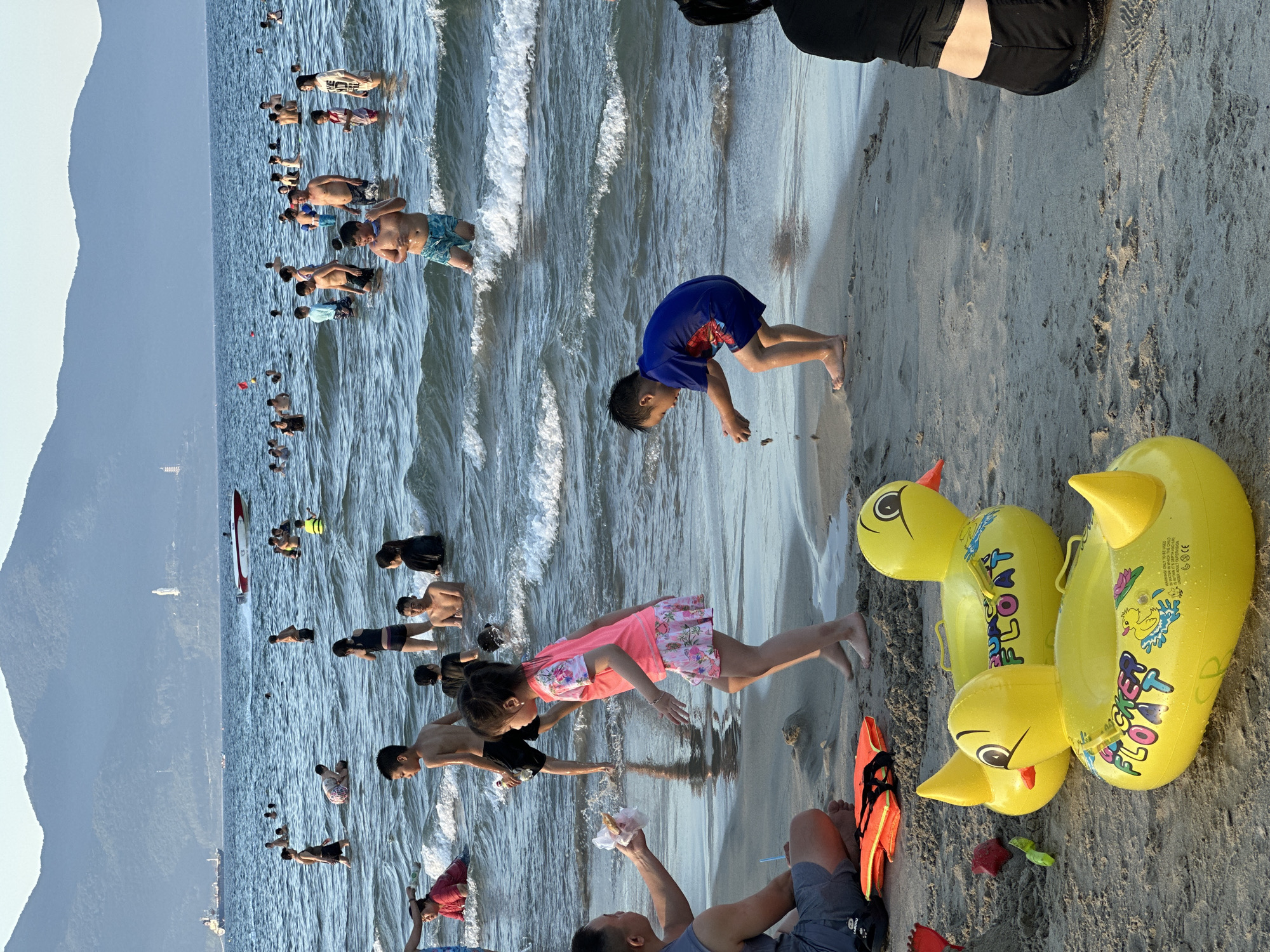 The image depicts a crowded beach scene at Da Nang, Vietnam, likely during the day. The sea is calm with gentle waves, and many people are in the water, some wading and others swimming. The beach is sandy, with footprints visible.
The subjects are predominantly people of various ages enjoying a day at the beach. There are children playing in the water and on the sand. In the foreground, two large inflatable yellow ducks are placed on the sand, along with an orange life vest. One of the ducks has text that reads "DUCKET FLOAT" and possibly other details related to flotation devices.
The activity is leisure and recreation. People are cooling off in the sea, playing in the waves, and relaxing on the beach. The overall atmosphere appears to be one of enjoyment and fun.
Notable details include the clear, calm sea and the presence of a distant mountain range on the left side of the image, which is a characteristic of the Da Nang coastline. The lighting suggests it is likely late afternoon, given the long shadows cast on the sand. The sky is clear and bright. The presence of the inflatable ducks and life vest indicates a family-friendly outing. Some individuals are wearing swimsuits, further emphasizing the beach setting. The overall mood seems happy and relaxed, with people engaging in typical beach activities.