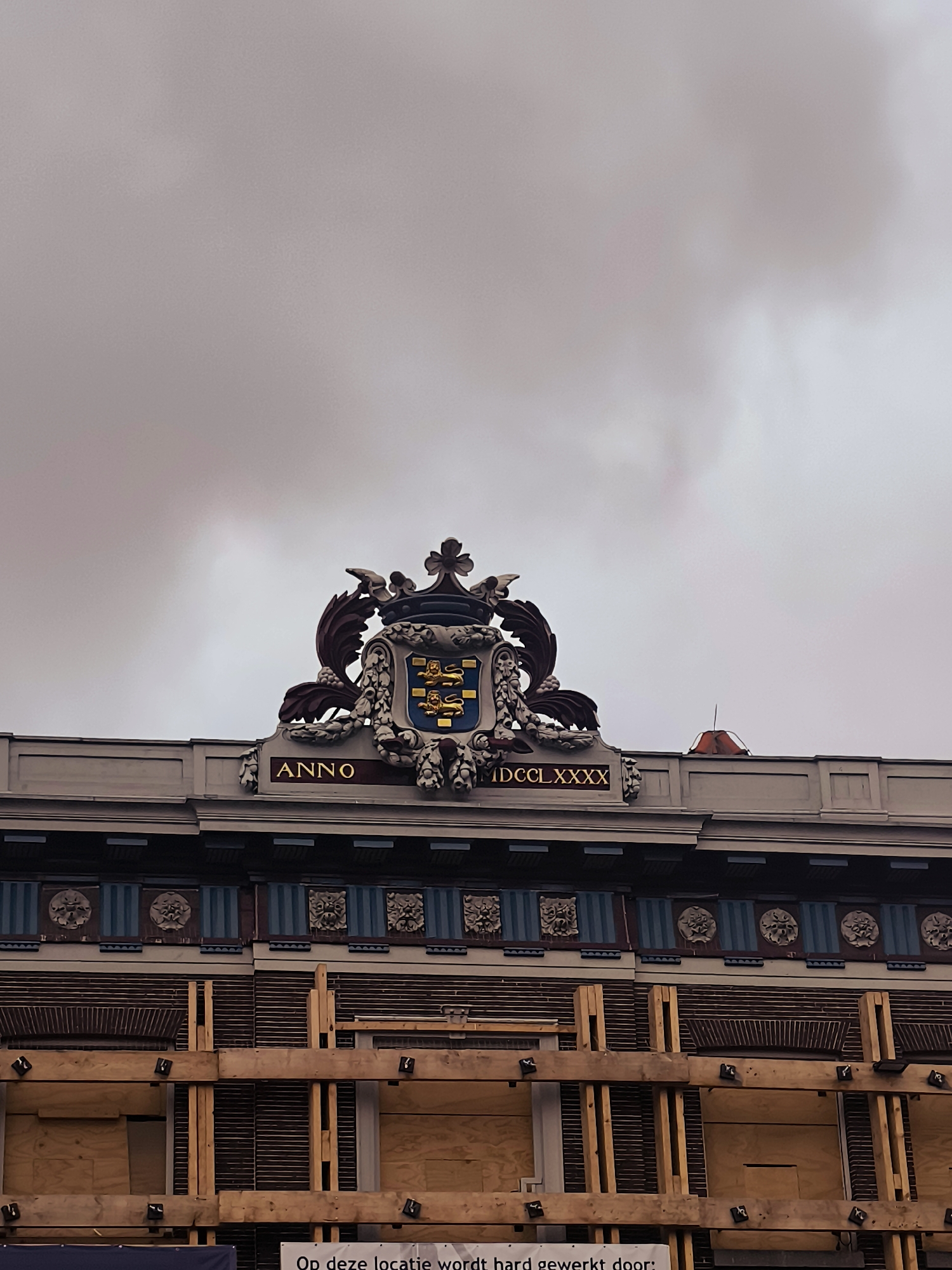 The image shows the facade of a building with ornate architectural details. At the top, a crest with a shield and two lions is prominently displayed. Below the crest, the words "ANNO" and "MDCCLXXXX" are visible in gold lettering, indicating the year 1780. The building's facade features decorative elements, including what appear to be carved rosettes or masks, set against a background of blue and dark stripes. The lower part of the building is under renovation or construction, with wooden scaffolding and plywood covering windows or openings. The sky above is overcast with grey clouds. The building is located in the city of Hoorn, Netherlands, at the address Grote Noord 84-90.
