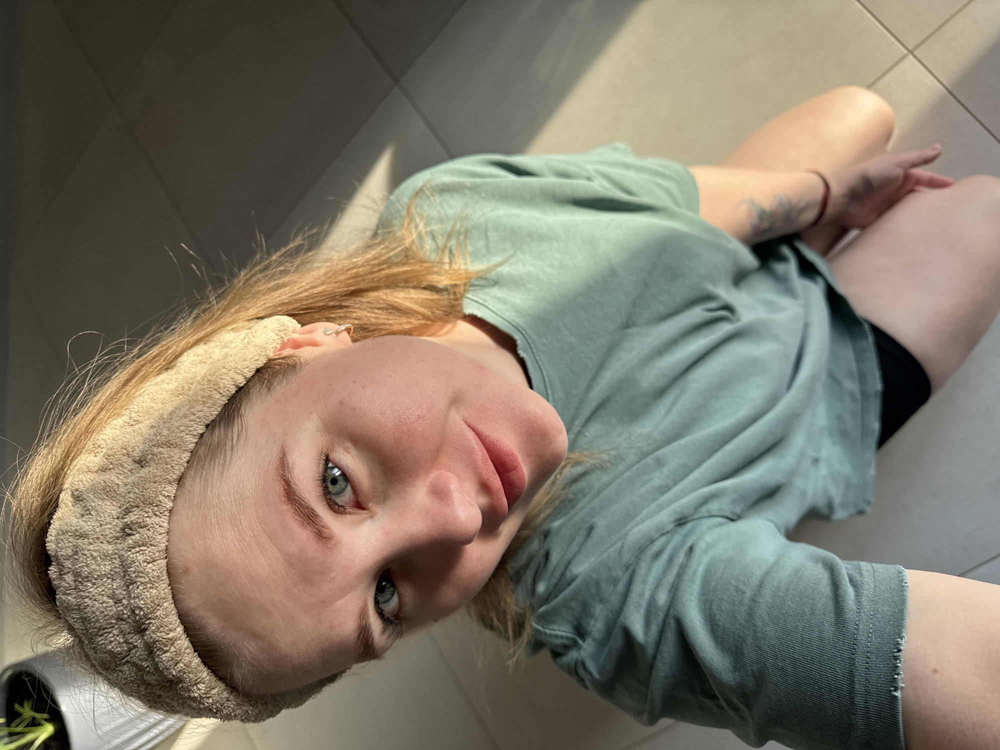 A person with light skin and light brown hair, wearing a beige headband and a sage-green t-shirt, is photographed from a slightly high angle. The person is sitting with their legs visible, and the background consists of light-colored tiles. A small plant pot with green sprouts is visible in the lower left corner of the image. The lighting is strong and casts shadows on the floor and the person's body. There is a tattoo on the person's left forearm. This image was not taken in the city of Gornyi (Dvurechenskoe S/P), Russia, as there is no discernible evidence in the image to suggest such a location.