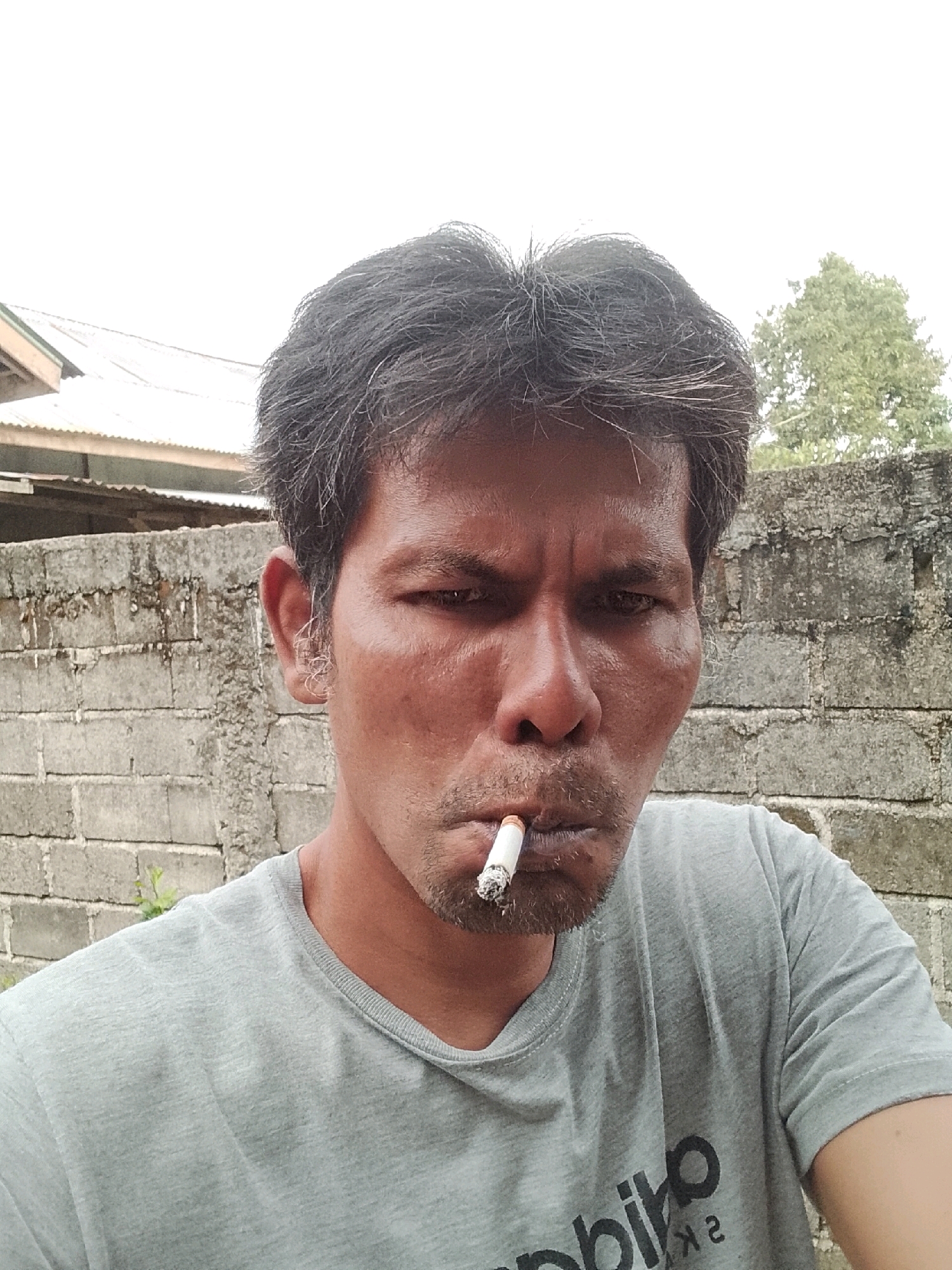 A man is captured in a close-up, selfie-style shot, his face filling the frame. He is smoking a cigarette, which is held between his lips, with the tip showing a faint glow. His eyes are narrowed, looking directly at the camera, and his expression is serious. The man has dark, somewhat disheveled hair, and stubble on his chin and upper lip. He is wearing a plain grey t-shirt.
The background consists of a rough, grey brick wall, weathered and possibly aged. Behind the wall, a glimpse of a building with a corrugated metal roof can be seen, suggesting an outdoor or semi-outdoor setting. A tree with green foliage is also visible in the distance, peeking over the wall. The overall lighting appears to be natural daylight, casting subtle shadows on the man's face and highlighting the textures of the brick wall. The scene suggests an informal moment, possibly in a residential area in the city of Padang Dalom, Indonesia.