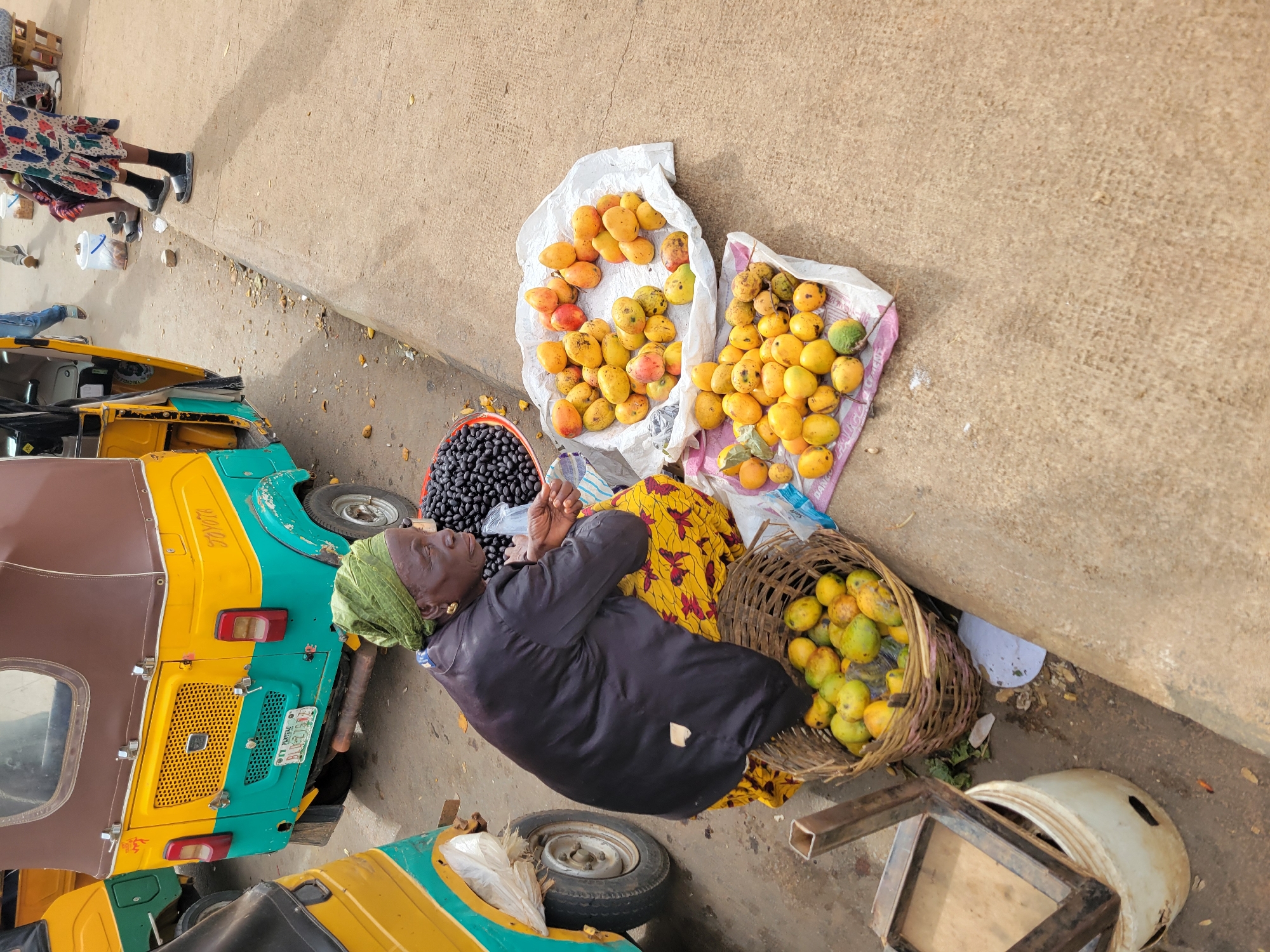 The image captures a street scene in Jos, Nigeria, where a woman is selling produce. She is seated on the ground next to two colorful auto-rickshaws, a yellow and green one prominently in the background and a partial view of another to the left. The woman, dressed in a dark top and a vibrant yellow and red patterned skirt, wears a green headscarf and gold earrings.
In front of her, on the ground, are two piles of mangoes displayed on white fabric, and a woven basket filled with more mangoes. To her left, a large round red basin overflows with dark berries, likely dates. Behind her, resting against the auto-rickshaw, is a large sack, possibly containing more goods. The ground is paved with a mix of dirt and concrete, showing scattered debris. In the distance, other individuals can be seen, and a wooden stool is visible, suggesting a bustling market environment. The overall scene depicts a typical day at a local marketplace in Jos.