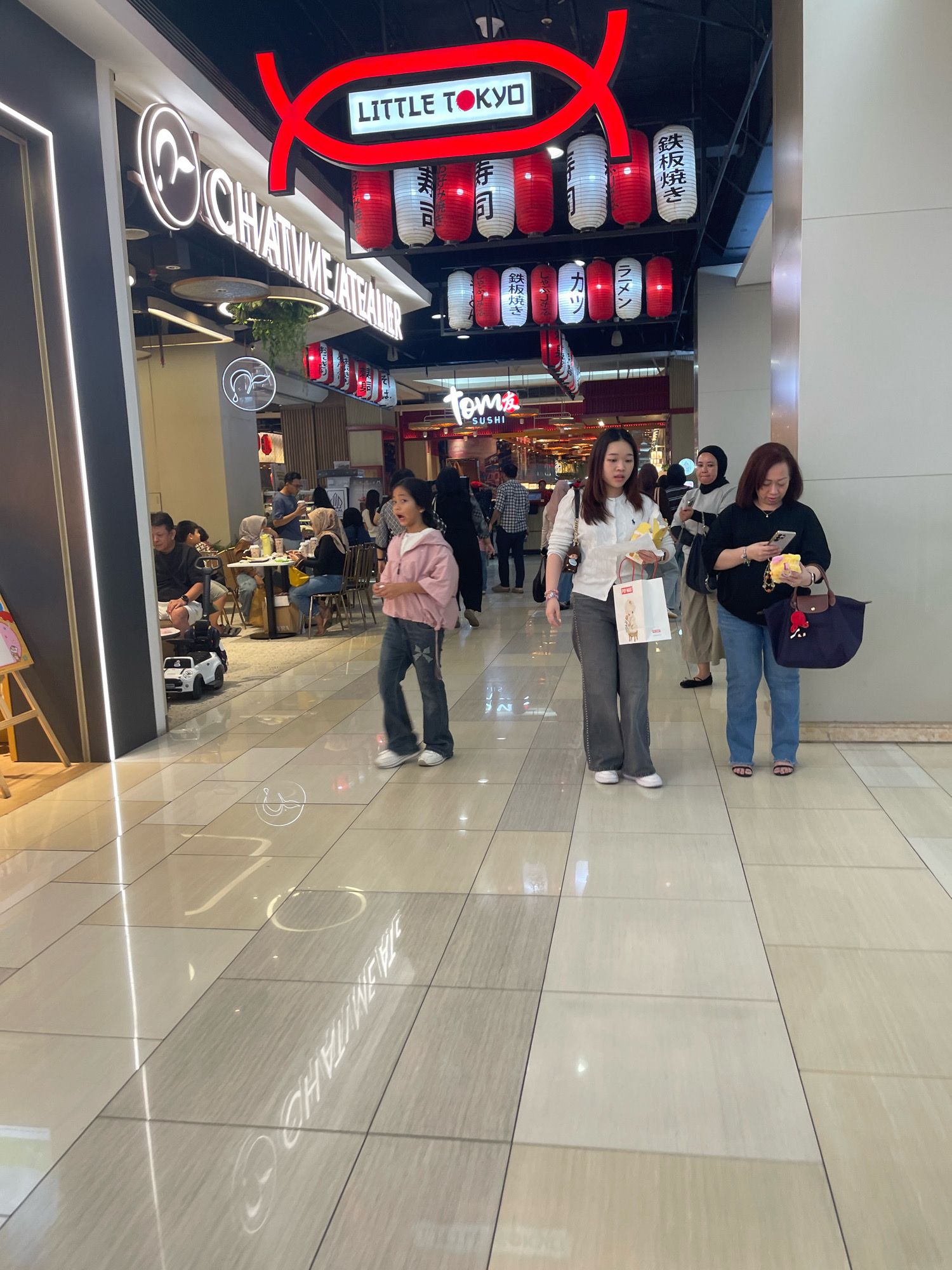 The image depicts a scene inside a shopping mall in South Jakarta, Indonesia. In the foreground, a young person wearing a pink jacket and jeans is walking, looking towards the right with an open mouth. To the right of this individual, two women are walking. One, in a white top and grey pants, is carrying a white shopping bag and a yellow item. The other woman, in a black top and blue jeans, is holding a phone and a small yellow object, with a large navy blue tote bag on her right arm. In the background, several people are seated at tables in what appears to be a restaurant area. Above the restaurant, there are signs for "LITTLE TOKYO" and "CHATIMEATEA LEAUER". Japanese-style lanterns, red and white, are hung as decoration. The flooring is tiled with a reflective surface. The lighting is bright, indicative of an indoor commercial space.