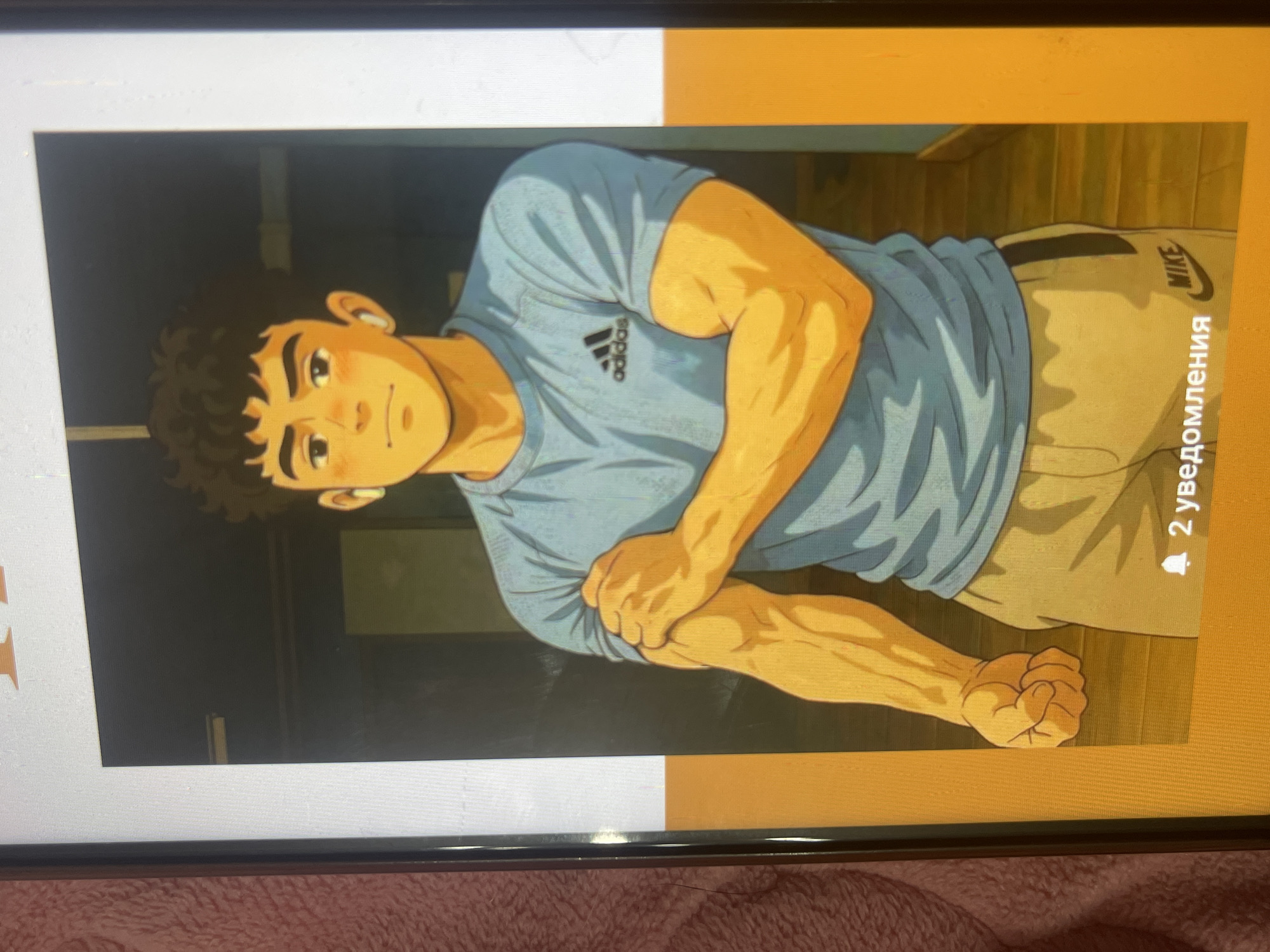 The image is a digital illustration of a young man in an anime style. He has dark, curly hair, fair skin, and is wearing a light blue Adidas t-shirt and tan Nike sweatpants. He is flexing his bicep. The background appears to be an indoor setting, possibly a room with a wooden floor and a doorway. A notification icon with Russian text is visible in the bottom right corner of the image. The provided image does not contain any information that would allow for its location to be determined in Bishkek City, Kyrgyzstan.