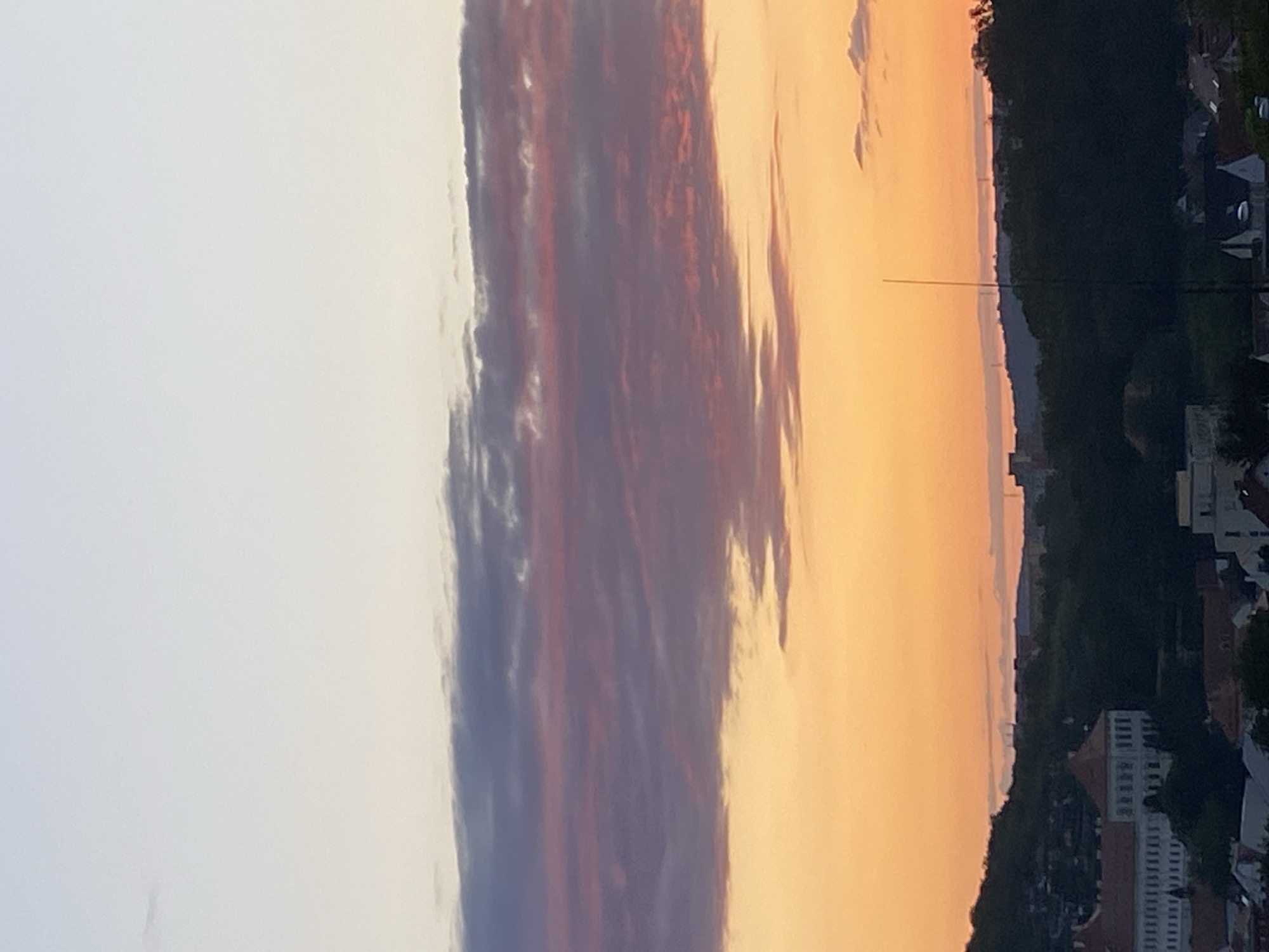 The image captures a dusky sky over the city of Bílina, Czechia. The sky transitions from a pale blue at the top to vibrant orange and pink hues as it nears the horizon, indicative of either sunrise or sunset. Wispy clouds, tinged with purple and pink, are scattered across the sky. Below the sky, a dark silhouette of trees and hills forms the backdrop of the city. In the foreground, several buildings are visible, with a prominent large building on the left featuring a red-tiled roof and white facade. A tall, thin antenna-like structure rises vertically in the middle ground, breaking the skyline. The overall scene depicts a tranquil urban landscape under a colorful twilight sky.
