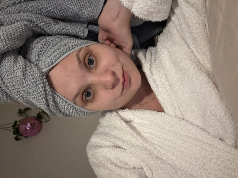 A close-up, indoor selfie depicts an adult female with light skin and blue eyes, looking directly at the camera. She wears a white terrycloth bathrobe and has a light gray, textured towel wrapped around her head like a turban. A thin gold-colored chain necklace is visible around her neck. Her left hand is partially visible, with fingers resting gently near her left ear/jawline. The background consists of a plain, light-colored wall. In the upper left quadrant, a hanging plant with green leaves is visible, adorned with a shiny, pink, spherical ornament. The lighting is soft and appears artificial. The specific location within Tel-Aviv, Israel, cannot be determined from the visual evidence provided in the image.