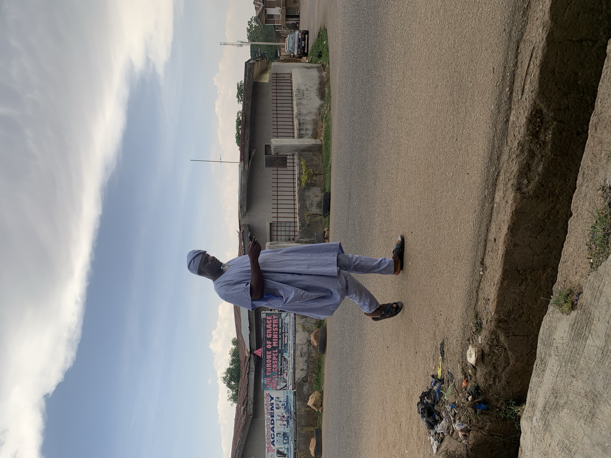 A man in a light blue traditional outfit stands on the roadside in Gura Top, Nigeria, looking at his phone. He is on the edge of a road next to a drainage ditch filled with litter. Behind him, there's a building with a sign advertising the "Precious Stone Academy" and "The Throne of Grace Int'l Gospel Ministry." The sky is partly cloudy.