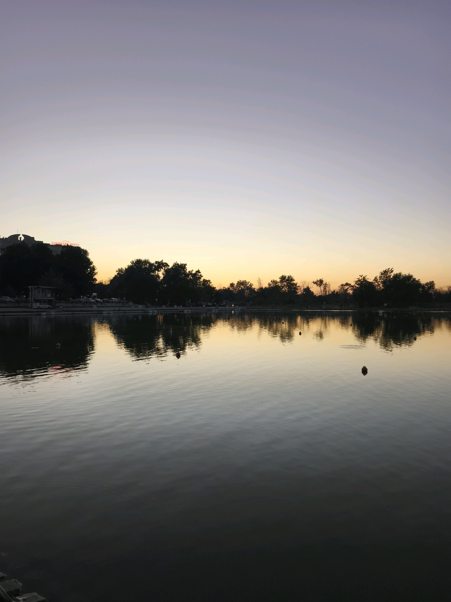 The image depicts a serene lake at dusk in an urban park. The sky transitions from a pale lavender at the top to a soft yellow near the horizon, indicating either sunrise or sunset. Silhouetted trees line the far bank of the lake, reflecting in the calm water. To the left, a building with illuminated signage reading "LANDMARK HOTEL" stands prominently, partially obscured by trees. Cars are parked along the shore. The water is smooth, with gentle ripples, and a few dark buoys are visible on its surface. The foreground shows the edge of the lake with what appear to be concrete steps or a retaining wall. The overall atmosphere is tranquil and peaceful, with the city lights beginning to subtly illuminate the scene.