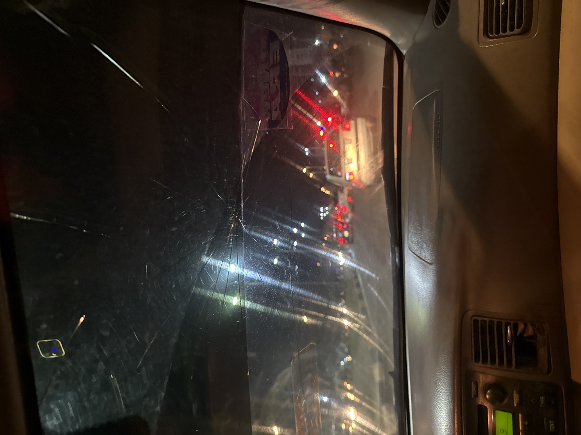 A view from inside a vehicle at night, presumably in Abuja, Nigeria, based on the presence of a vehicle permit sticker. The windshield is cracked, obscuring the view of traffic and streetlights. The dashboard and interior of the car are visible on the right side. The scene suggests the vehicle is stationary or moving slowly in heavy traffic.