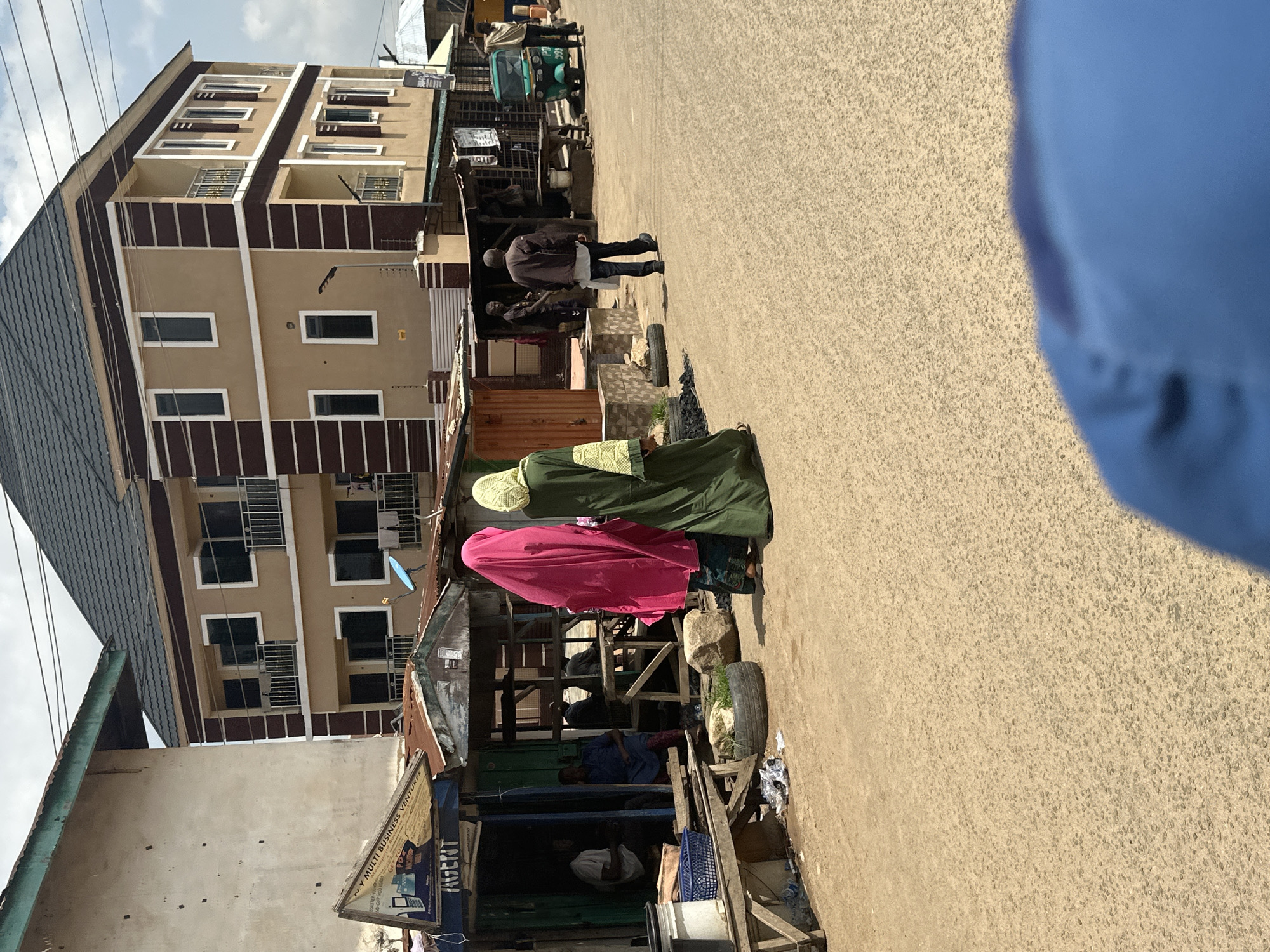 This image depicts a street scene in Jos, Nigeria. The primary subjects are several people, including two women in traditional Islamic attire, one wearing a bright pink hijab and the other a green abaya. They are standing in what appears to be a market or commercial area, possibly interacting with vendors.
The scene is set along a paved road with a multi-story building on the left. This building has a modern aesthetic with beige and brown accents, multiple windows with metal bars, and a dark-colored roof. Adjacent to the building, and in the foreground, are market stalls constructed with wood and corrugated metal, suggesting a bustling commercial environment. Some of these stalls have signs, though the text is mostly illegible. One visible sign appears to advertise "MULTI BUSINESS VENTURES" and possibly has contact information.
In the background, other individuals are visible, some walking along the road, and one man is seated under a stall. There is also a small motorized tricycle, common in this region, parked further down the street. The ground is a sandy, gravelly asphalt. The lighting suggests it is daytime, likely mid-morning or afternoon, with the sun casting shadows. The sky is partly cloudy.
The overall impression is one of everyday life in a busy urban or semi-urban area of Nigeria, with a blend of modern architecture and traditional market structures. The presence of people in traditional clothing and the type of vehicles suggest a cultural context specific to the region.