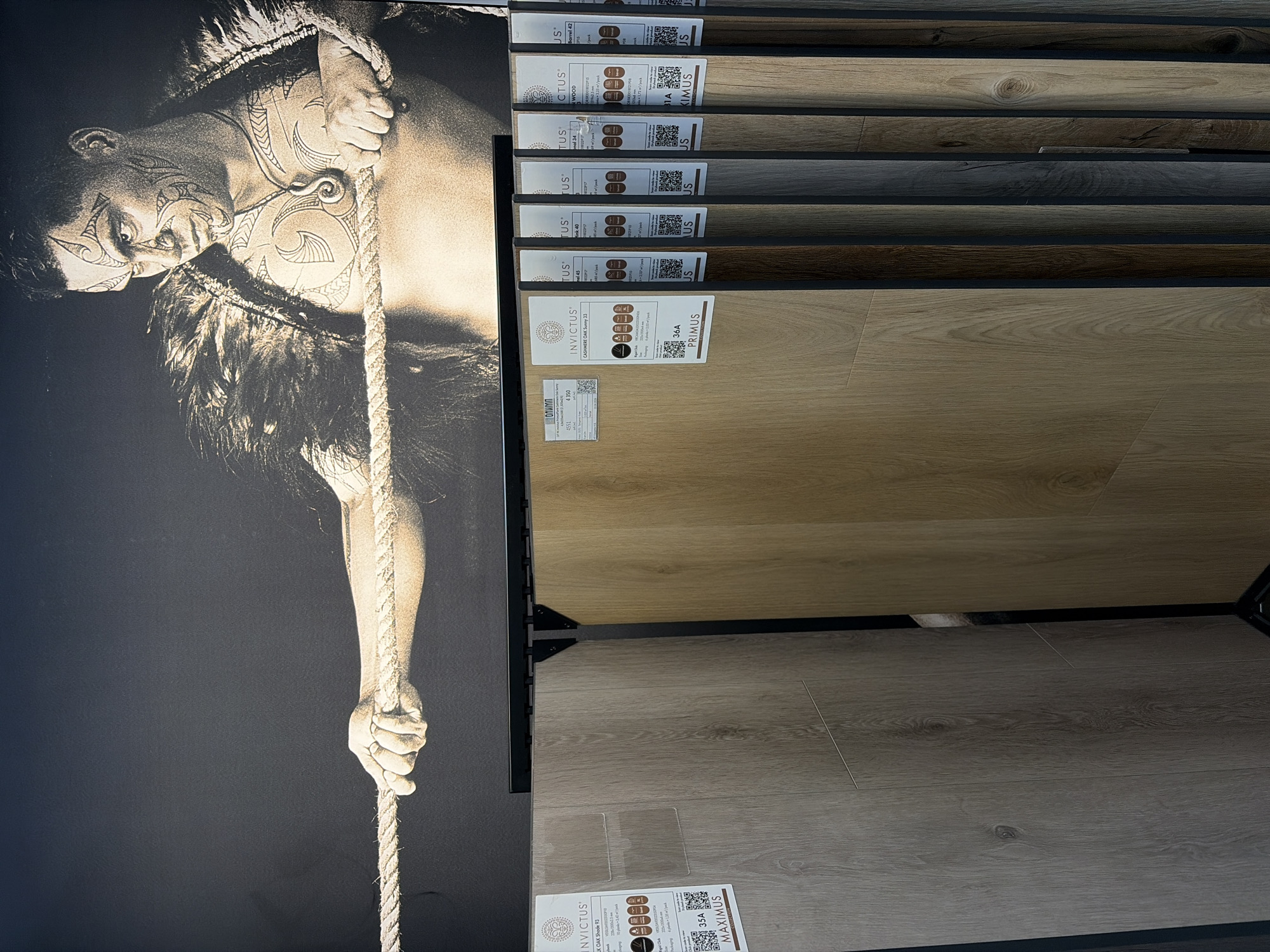 The image shows a vertical display of Invictus flooring samples. The samples are arranged in a vertical stack, varying in color and texture, from light to dark wood tones. Each sample has a label with text that reads "INVICTUS", name or shade number. To the left of the flooring samples, there is a black and white image of a man with tribal tattoos climbing a rope. In the bottom left corner, two labels are partly visible with text "MAXIMUS", "93", "35A".
The image provides no information about location.