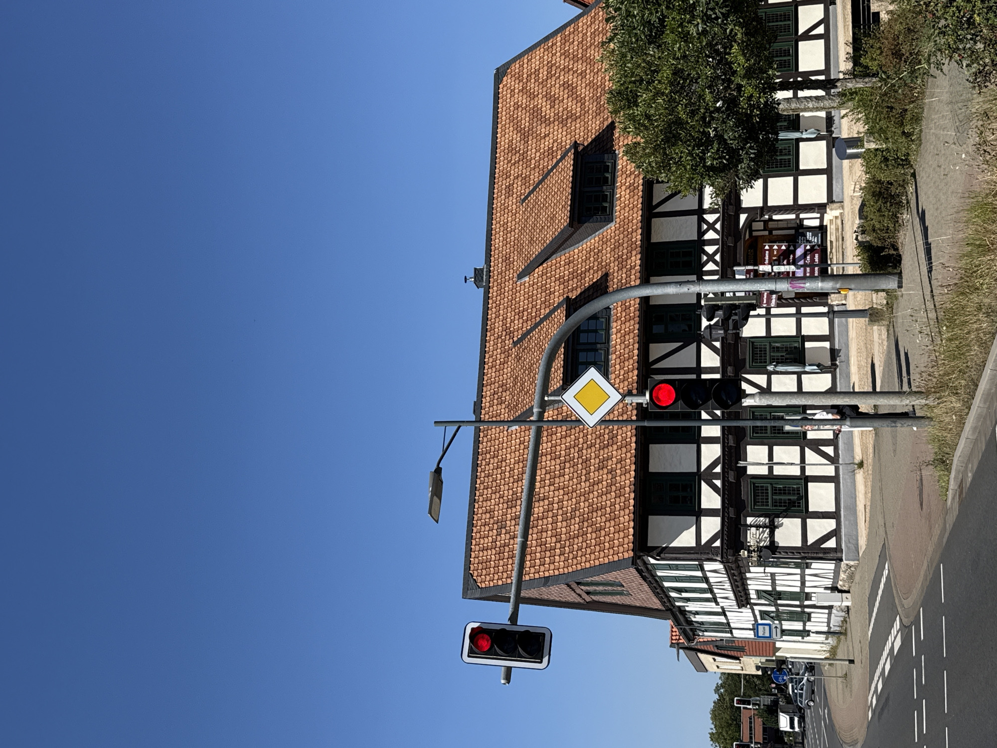 The image shows a clear blue sky over the town of Herzberg, Germany. In the foreground, a street with a pedestrian crossing is visible. To the right of the street, a building with a traditional half-timbered facade stands. The building has a red tiled roof and dark green shutters. In front of the building, there is a tree and some bushes. A traffic light with a red signal is positioned on the right side of the image. Another traffic light and a diamond-shaped yield sign are visible on the left side of the image. The overall scene is a street view in a German town, likely during daytime due to the bright sunlight and clear sky.