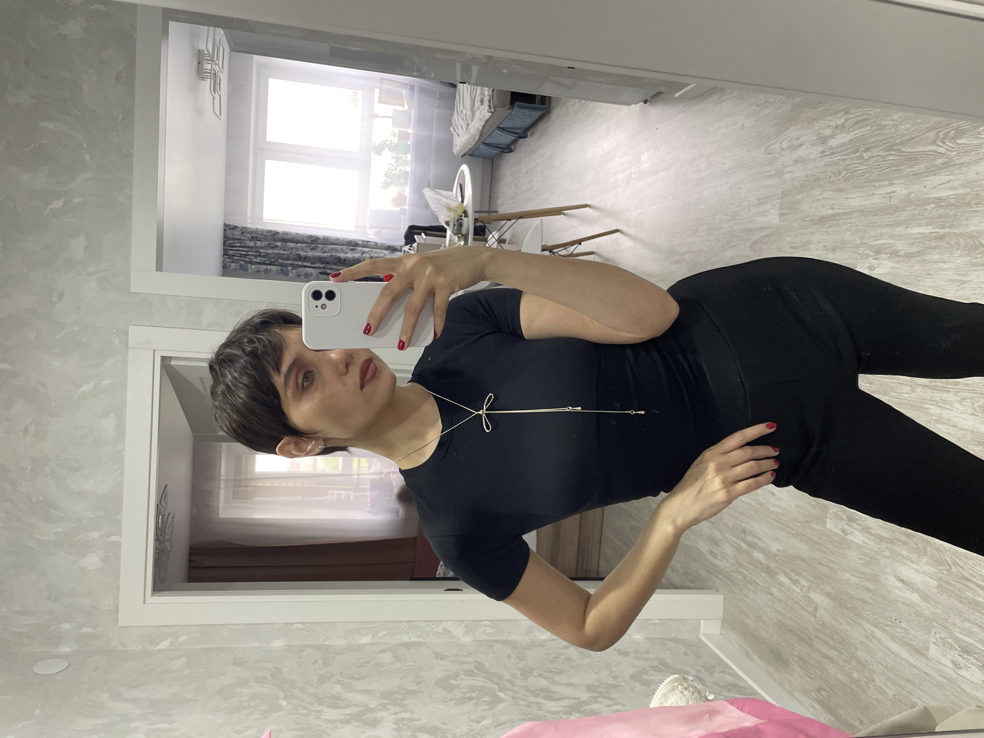 A woman is taking a selfie in a mirror, likely in a home setting. She's wearing a black top and leggings. Her hair is short and dark. She is holding a white smartphone with red nail polish on her nails. The background includes a glimpse of an interior with a window, curtains, furniture, and a white door frame. The precise location in the city of Izhevsk, Russia, cannot be determined from this image alone.