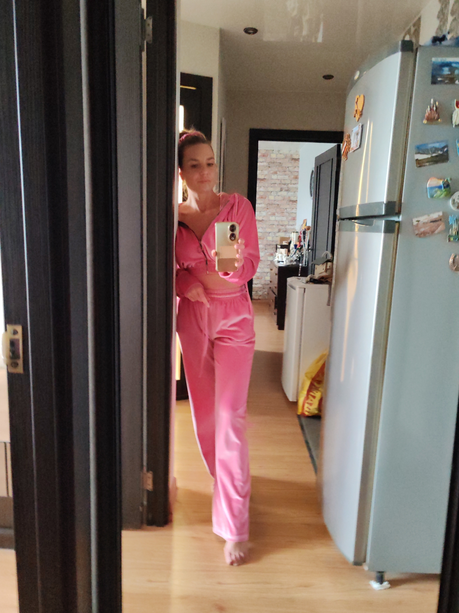 A woman is visible in a mirror, standing in what appears to be a kitchen or hallway. She is wearing a pink velour tracksuit and holding a cell phone. The room features light-colored wooden flooring, a refrigerator covered in magnets, and a doorway leading to another room with exposed brick and furnishings. The location cannot be determined to be Petropavlovsk-Kamchatsky, Russia from the image alone.