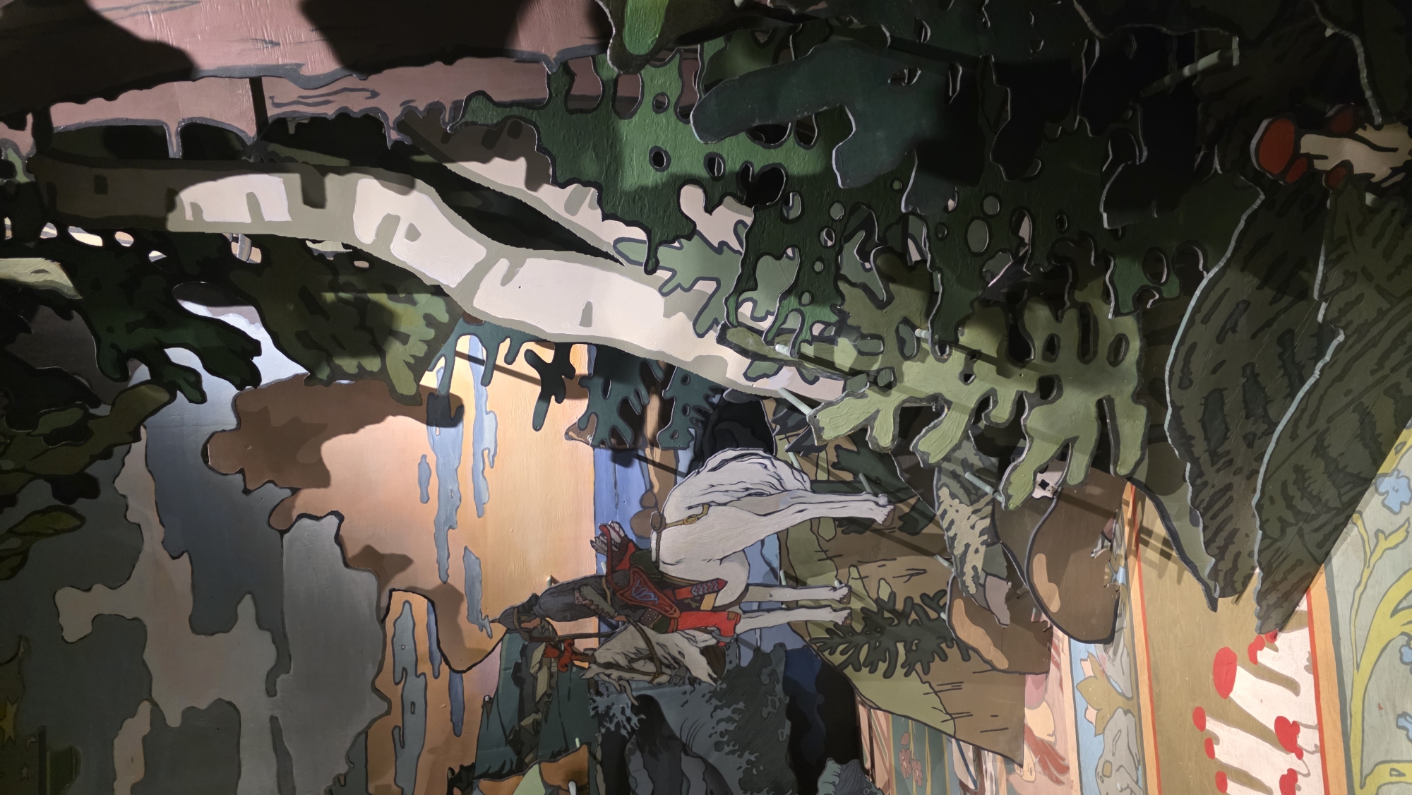 The image depicts a multi-layered diorama or theatrical set design. The foreground features painted cutouts representing trees with green and gray foliage. Behind the trees is a depiction of a scene with a person in red attire riding a white horse. This scene is layered against a background of abstract shapes and colors, including peach, blue, and gray. Some areas of the background show a more detailed floral design. The location is the St. Petersburg Puppet Theater, Russia.
