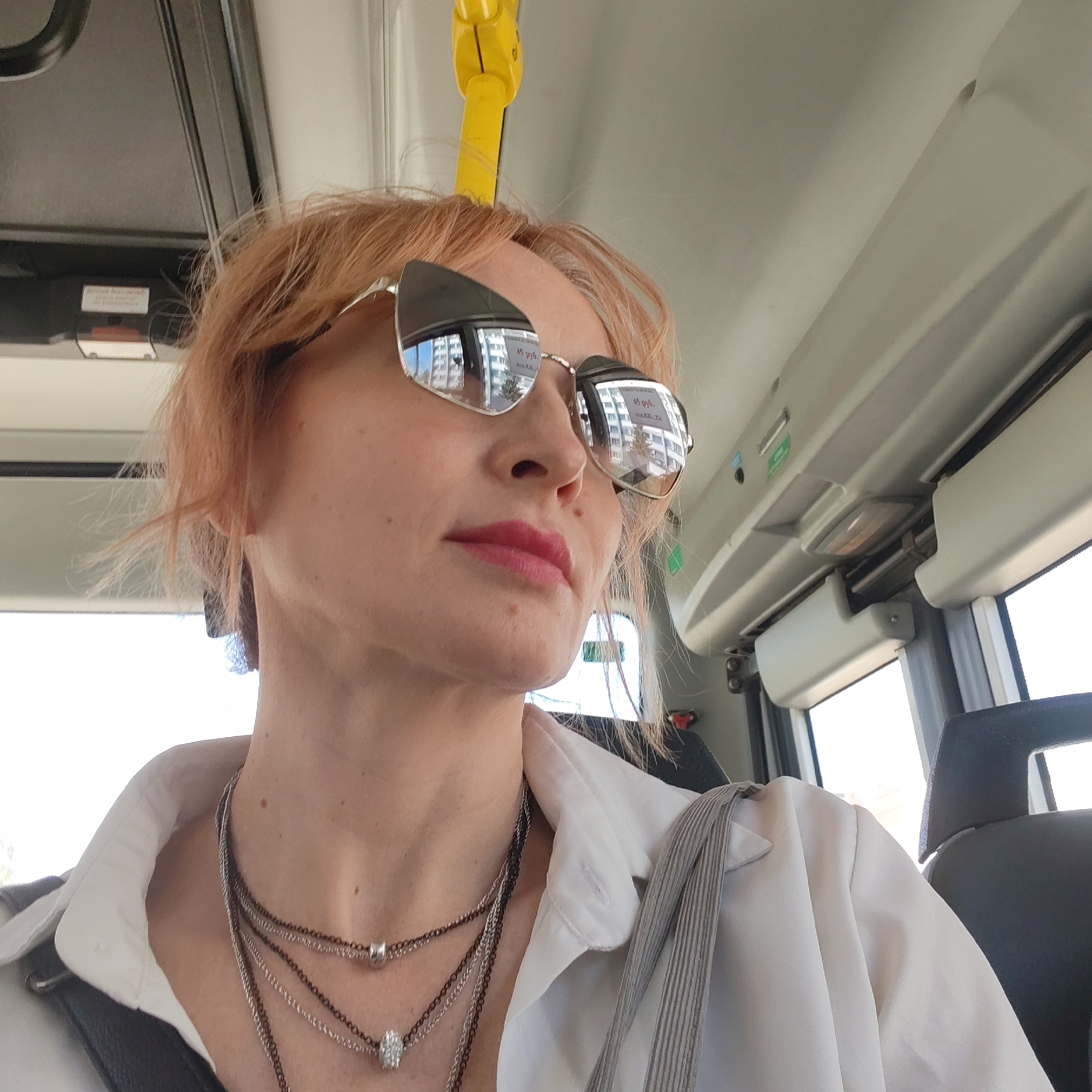 A light-skinned woman with short, light auburn hair is sitting on a bus. She is wearing sunglasses that reflect a city scene with buildings and trees, and her lips are painted a pinkish-red color. She is wearing a white button-down shirt and several necklaces. A yellow handrail is visible above her head, indicating she is likely riding public transportation. The interior of the bus is visible, including windows and seating. According to the reflection in the sunglasses, the woman is in the city of Chaikovsky, Russia. The buildings reflected in the sunglasses and the interior of the bus are essential environmental details.
