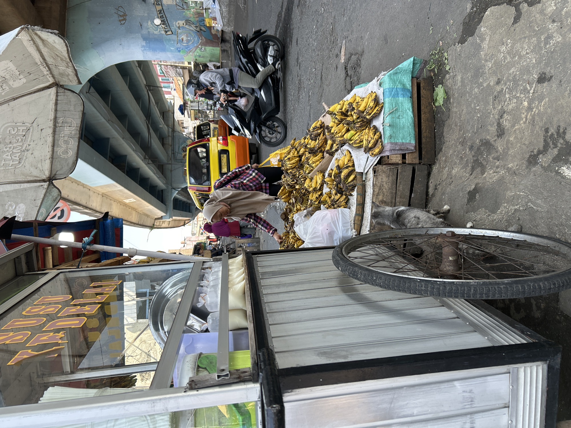 This image depicts a street scene in Depok, Indonesia, beneath an elevated highway structure. In the foreground, a silver food cart is visible, with its side and a large wheel prominent. The cart appears to be selling drinks in plastic bags, with some bottles of green and yellowish liquid inside. To the right of the cart, a vendor, a woman wearing a headscarf and a checkered shirt, is selecting bananas from a large pile. These bananas are displayed on a table covered with a green and blue cloth, placed on wooden crates. A gray cat is also present near the wheel of the cart. In the background, several vehicles are visible, including a yellow minivan and a black motorcycle with a rider wearing a helmet. The overall environment suggests a busy, outdoor market or street-side vendor area.