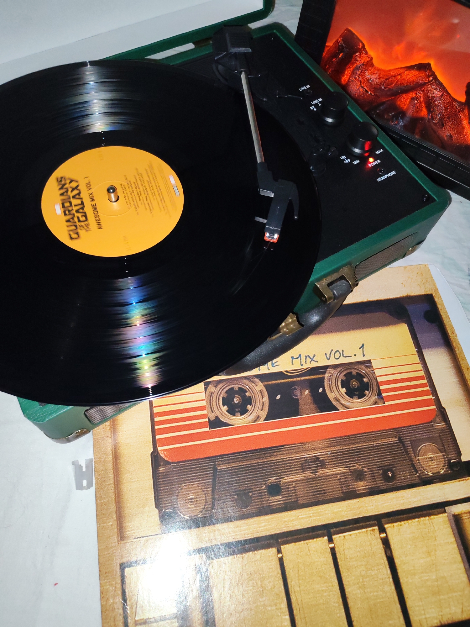 An overhead view captures a dark green portable record player actively playing a black vinyl record. The record's yellow label prominently displays "GUARDIANS OF THE GALAXY AWESOME MIX VOL. 1" in black lettering, with "I'M NOT IN LOVE" visible among the partially obscured tracklist. The tonearm, fitted with a red stylus cartridge, is positioned on the record, which reflects iridescent colors from ambient lighting.
The record player features a control panel with two black knobs and a red illuminated "POWER" light. Visible text on the panel includes "LINE IN," "LINE IN BT," and "HEADPHONE," along with "ON OFF MIN MAX" around one of the knobs. Beneath the record player, a record sleeve or art print depicts a vintage cassette tape with the text "THE MIX VOL.1." In the upper right background, a device with an orange-red glow, resembling a fireplace or embers, adds a warm ambiance to the scene. The setting appears to be indoors, possibly during the evening or night. While not visually discernible from the image, the context provided states the location as Izhevsk, Russia.