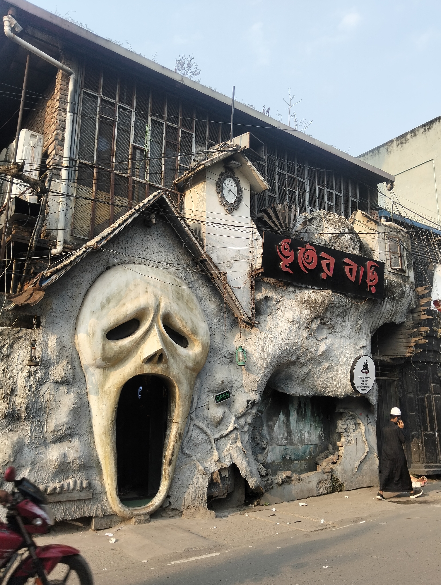 The image shows the entrance of "Bhuter Bari" (ভূতের বাড়ি), a haunted house attraction in Dhaka, Bangladesh. The entrance is designed as a large, distressed white face with the open mouth serving as the doorway. To the right of the face, the structure transitions into a cave-like appearance with a sign bearing the name in Bengali and a smaller, circular sign indicating that the attraction has no other branches in Lalbagh and warning against fraud. A man dressed in a black robe and a white cap is walking past the entrance, engaged in a phone call. A partially visible red motorcycle is parked on the left side of the frame. Overhead, the building is an aged two-story structure with barred windows and a clock tower detail. Wires are visible along the façade. The scene is on a street with a curb and what appears to be a paved surface.