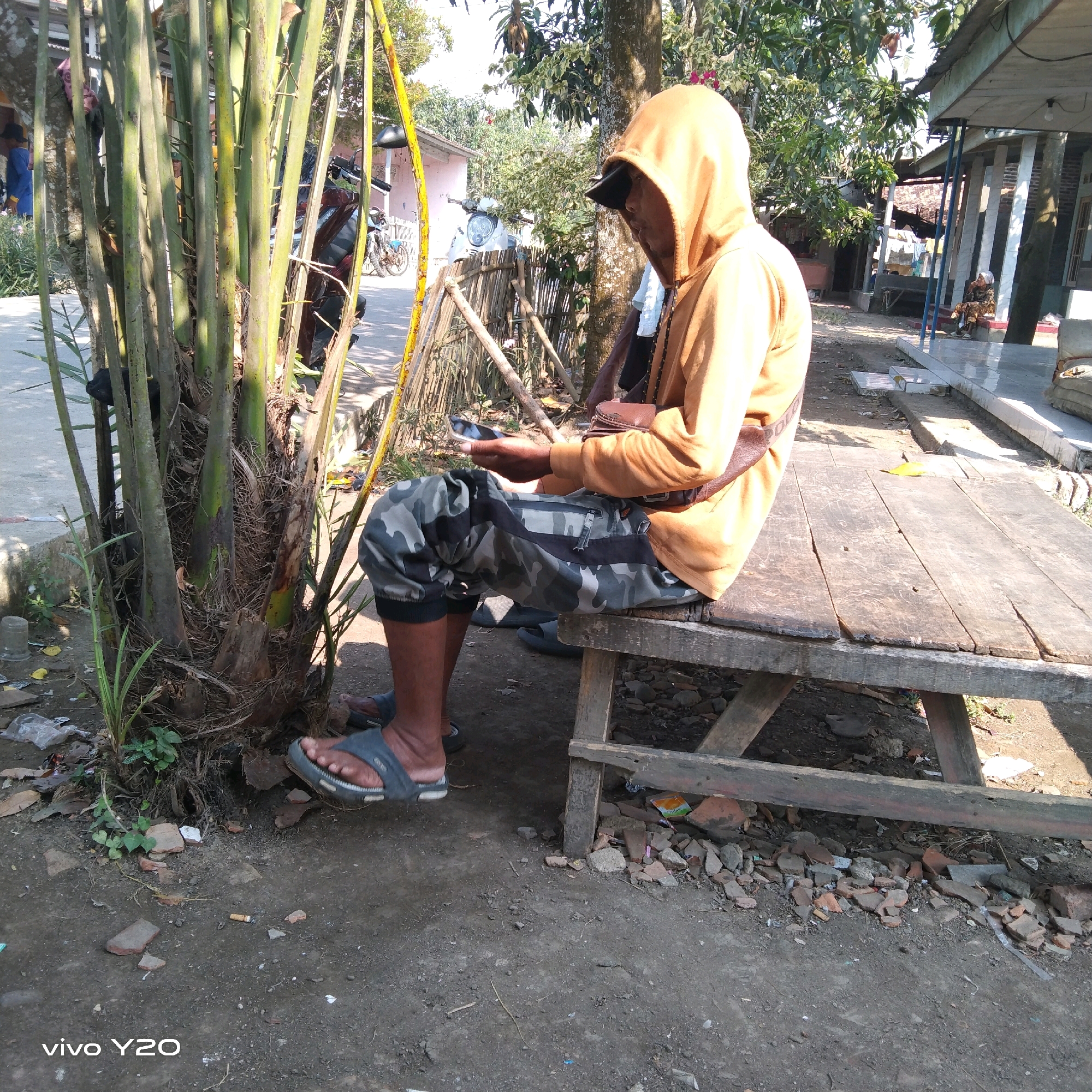 A person wearing an orange hoodie and camouflage pants sits on a wooden bench, holding a mobile phone. They are positioned in front of a cluster of tall, thin plants. The scene appears to be outdoors in a somewhat rural or unkempt environment, with dirt, scattered debris including broken bricks, and vegetation. In the background, there are buildings, a fence made of bamboo, and what appears to be a motorcycle. The lighting suggests it is daytime with sunlight casting shadows. The image is a close-up focusing on the person and their immediate surroundings.
