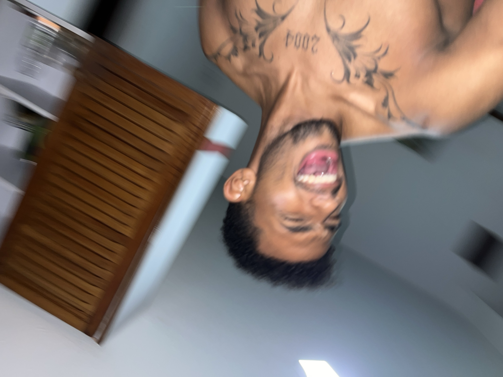 The image displays a young adult male indoors, captured in a dynamic, rotated composition with notable motion blur. The subject has dark hair and a beard, with his mouth open wide, revealing teeth and tongue, suggesting he is mid-laugh or shout. His eyes are squinted. He is shirtless, exposing several tattoos on his left upper torso: the numbers "2004" are visible on his left pectoral, accompanied by swirling, dark, ornamental or tribal-style designs extending across his shoulder and chest. The background features light-colored walls, with a distinct brown wooden structure composed of parallel slats visible in the upper portion of the frame, possibly a blind, shutter, or furniture panel. A bright, localized light source is present on the lower left, creating a glare. There are no specific visual cues within the image to identify its precise location within Porlamar, Venezuela; it depicts an interior, likely residential, setting.