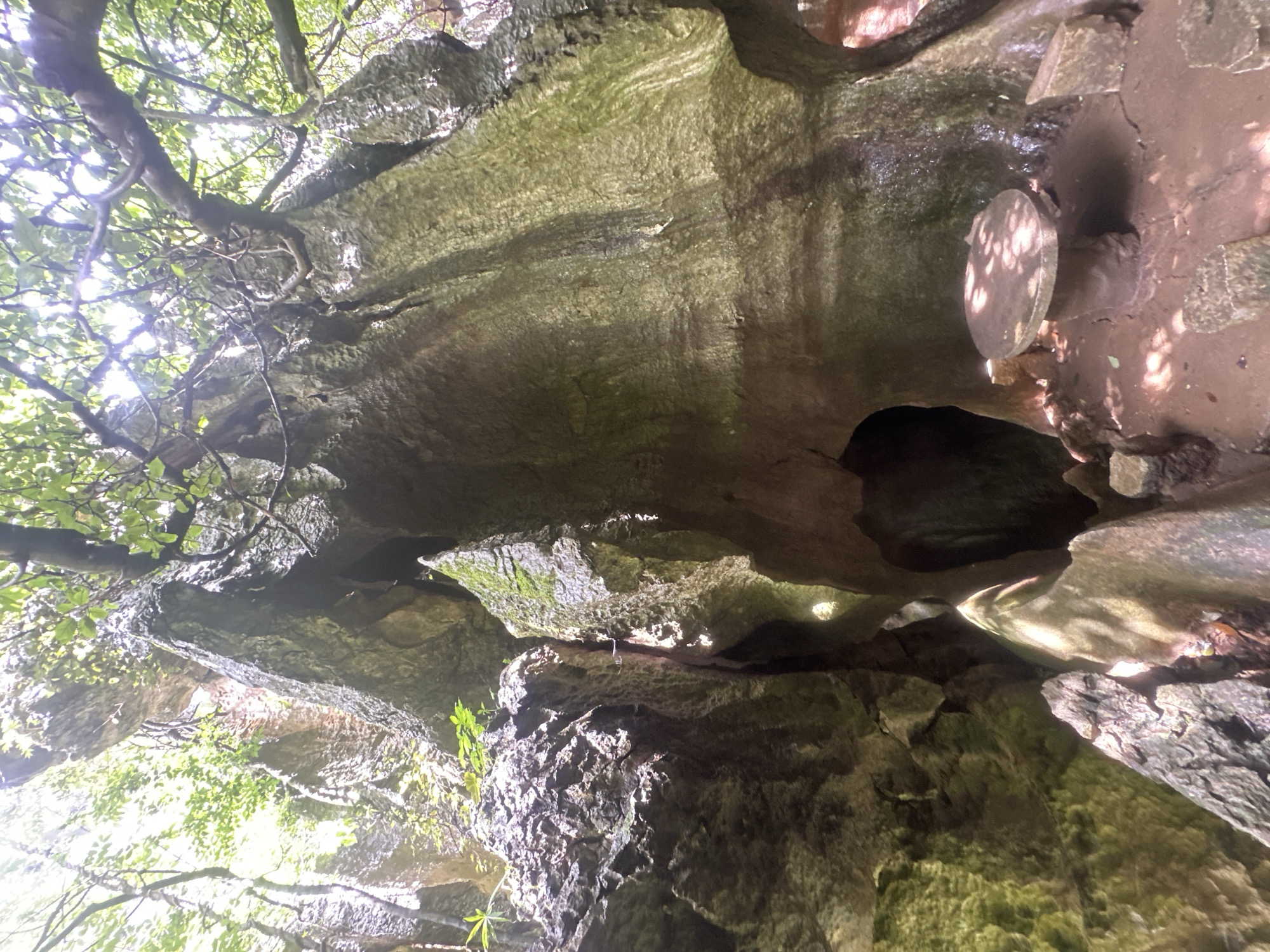 The image depicts a natural outdoor scene within the city of Kunming, China, likely in a karst landscape. The central focus is a large, weathered rock formation with a cave-like opening. To the right of the opening, a stone table with a circular top and a pedestal base is situated on the ground. The surrounding environment is characterized by rugged, moss-covered rocks and abundant foliage, with tree branches overhead creating a dappled light effect. The ground appears to be a mix of soil and smaller stones. The overall impression is of a secluded, natural spot.