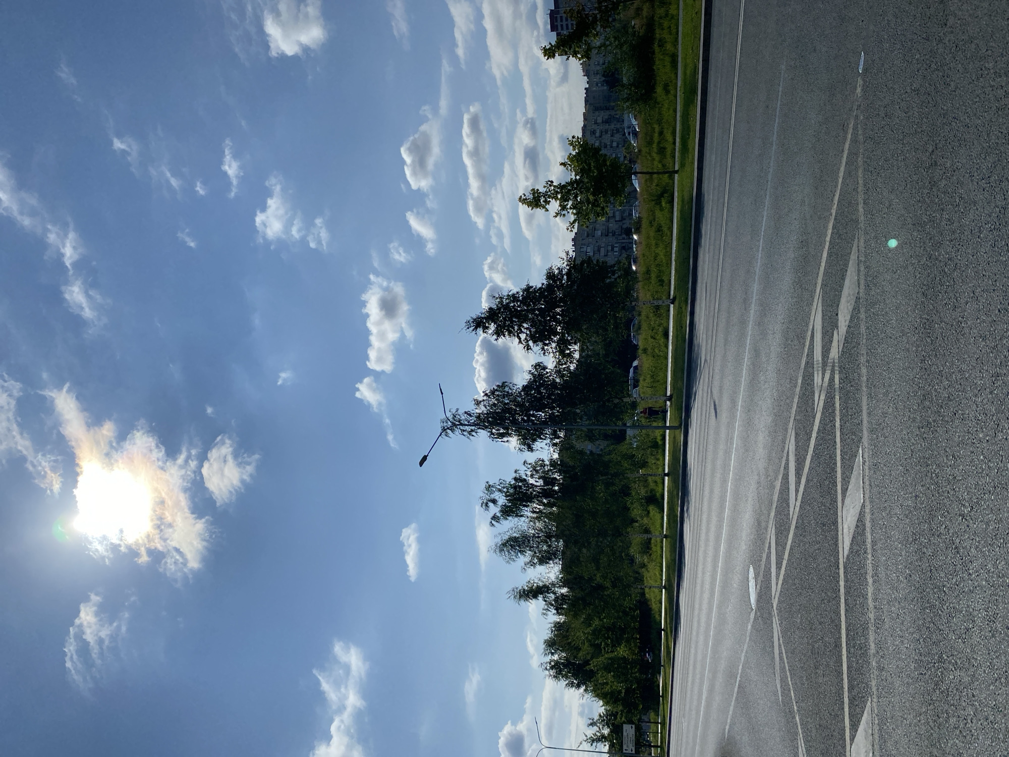 The image depicts a street scene in Saint Petersburg, Russia, on a sunny day with scattered clouds. A paved road occupies the foreground, with lane markings and a manhole cover visible. Beyond the road, a strip of green grass and trees lines the background. In the middle ground, several deciduous trees stand tall, some with visible white bark, and behind them, a row of apartment buildings stretches across the horizon. The sky is blue with white, fluffy clouds, and the sun is bright, partially obscured by a cloud. A few cars are parked along the street in the distance. A lamppost is visible on the right side of the image, and a person in red clothing is walking on the sidewalk. Another lamppost is visible on the far left.