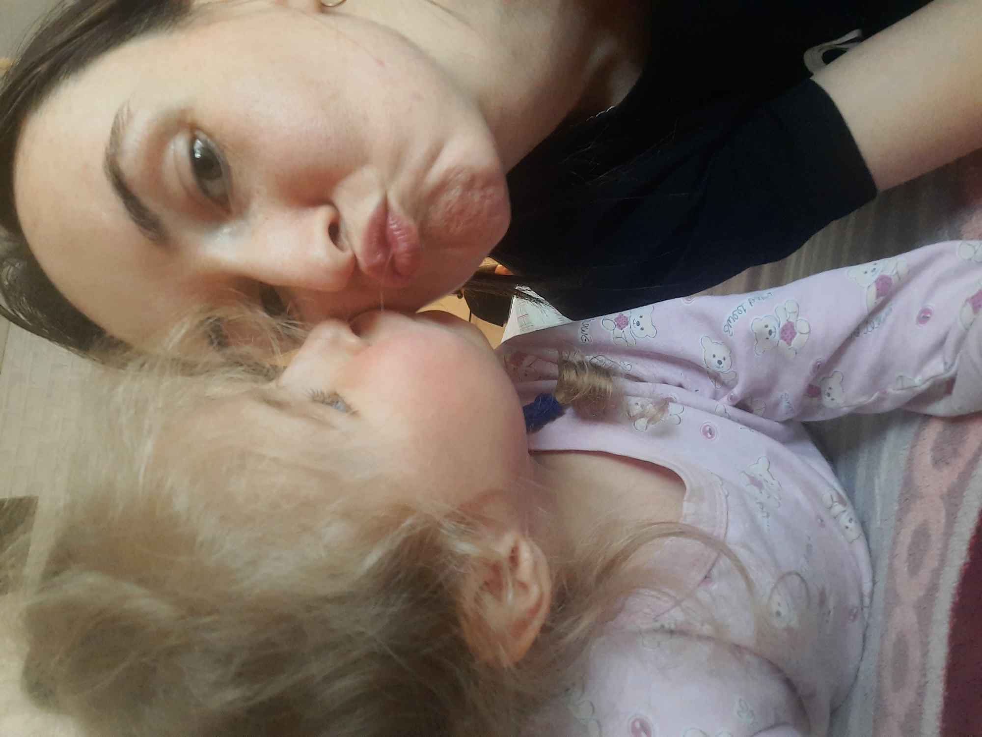 A woman and a young child are close together in an indoor setting. The woman has dark hair and is wearing a black top. She has her lips pursed as if she is kissing the child. The child has blond hair and is wearing a pink patterned long-sleeved pajama top. The child appears to be looking up at the woman. The background consists of a patterned surface, possibly a blanket or bedding. The location is Krasnogorskoye, Russia.