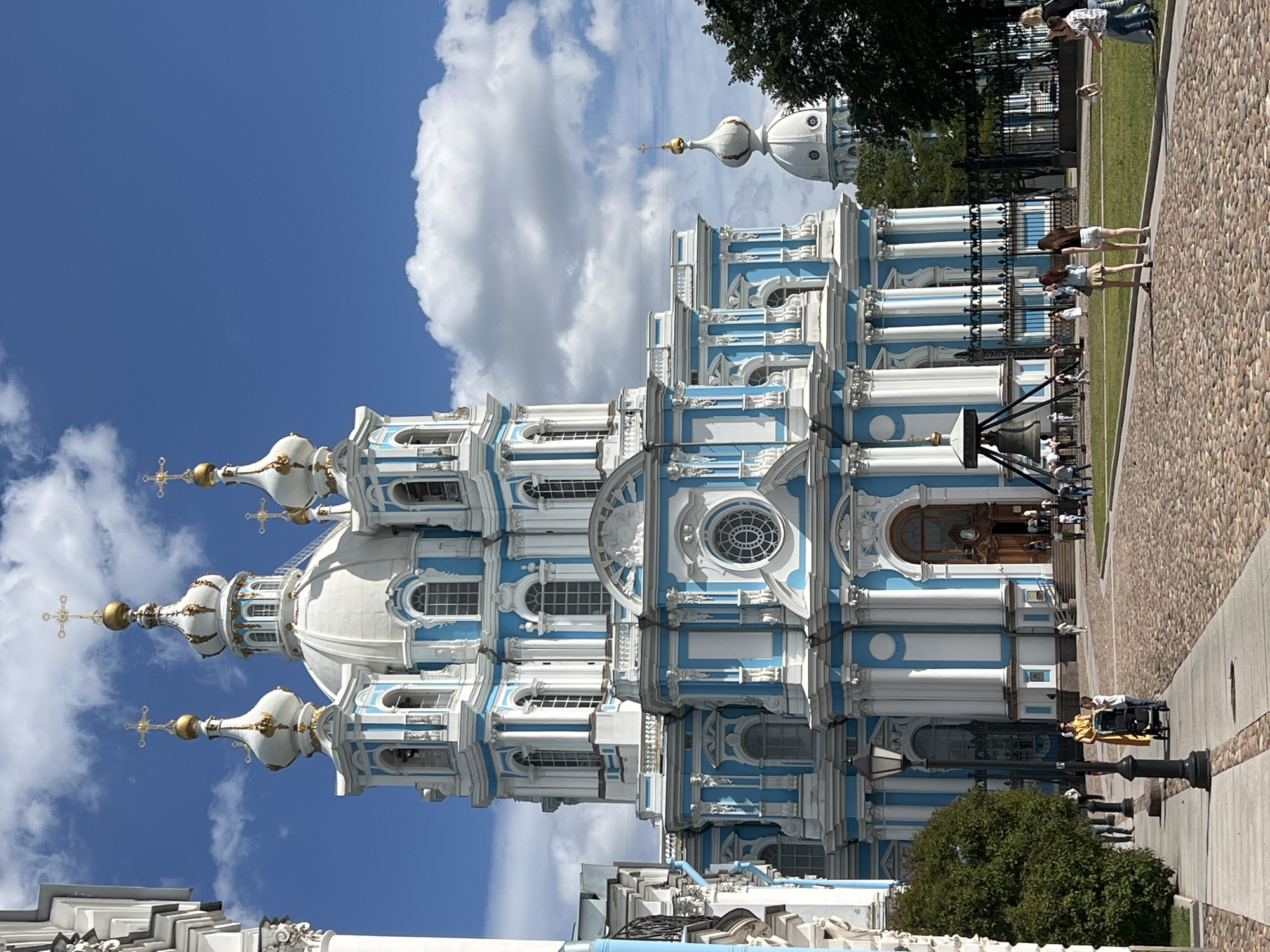 This is a sunny day image of the Smolny Cathedral in Saint Petersburg, Russia. The cathedral is a large, ornate blue and white building with multiple domes and golden crosses. In the foreground, a cobblestone path leads up to the cathedral entrance. Several people are walking on the path and on the grass in front of the cathedral, some are pushing strollers, and others are gathered in groups. A large bell is visible on the left side of the image. Lush green trees are present on the right side of the frame. The sky is a vibrant blue with fluffy white clouds.