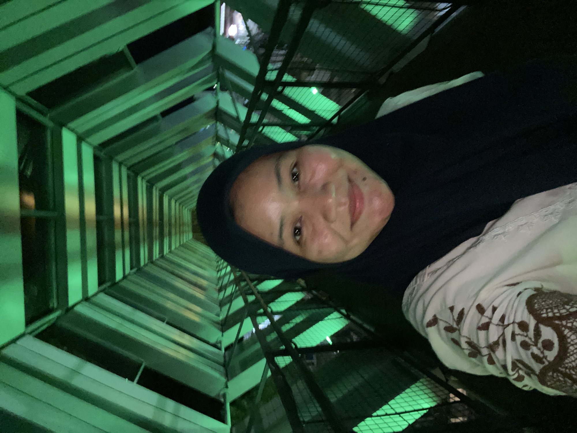 A woman wearing a black hijab and a light-colored shirt with brown floral embroidery takes a selfie in a tunnel-like structure with green lighting. The structure appears to be a pedestrian bridge or walkway, with a geometric design of angled beams and panels illuminated by green lights. The woman is smiling at the camera, and her face is partially visible. The background shows the repeating pattern of the bridge's architecture receding into the distance. The location is identified as the Banjarbaru City Bridge in Banjarbaru, Indonesia.