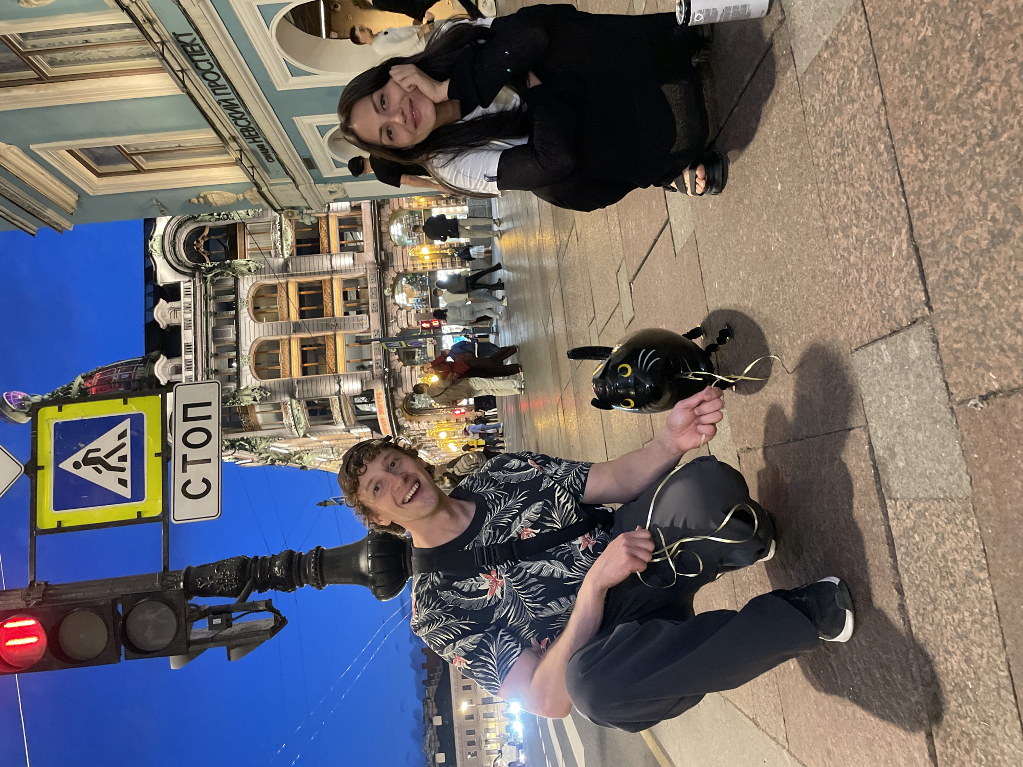 The image depicts an urban street scene at dusk or early night in Saint Petersburg, Russia. The location is identifiable by a building sign displaying "НЕВСКИЙ ПРОСПЕКТ" (Nevsky Prospekt) in Cyrillic on a light blue building on the right, and a "СТОП" (STOP) sign, also in Cyrillic, mounted on a street pole.
In the foreground, two individuals are squatting on a paved sidewalk. On the left, a man with curly brown hair and a dark baseball cap worn backward is smiling directly at the camera. He wears a dark short-sleeved shirt with a tropical leaf pattern, dark pants, and black athletic shoes. He holds several strands of golden ribbon in both hands. Between the man and the woman, a black cat-shaped balloon with yellow eyes and white whiskers lies partially deflated on the ground; the golden ribbons held by the man are connected to this balloon. On the right, a woman with long dark hair is also smiling at the camera, resting her chin on her left hand. She is dressed in a white t-shirt layered over a long-sleeved black top, black pants, and dark sandals. A white cylindrical object is visible on the ground near her right foot.
In the middle ground, a black traffic light pole features an illuminated red light, a blue square pedestrian crossing sign with a white walking figure, and the "СТОП" sign below it. The background consists of several multi-story historic buildings, illuminated against the twilight blue sky. The prominent light blue building on the right side of the street features ornate windows and arches. Further down the street, more blurred pedestrians are visible walking on the sidewalk. The pavement beneath the subjects is composed of rectangular granite slabs in reddish-brown and grey tones.