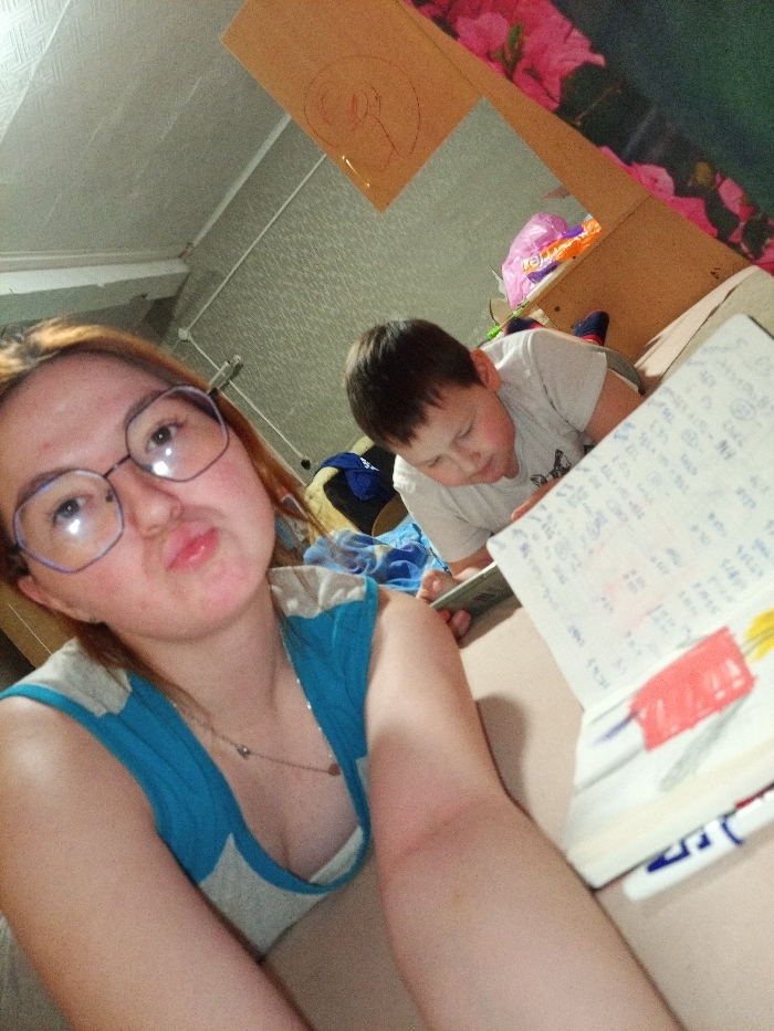 An indoor scene in what appears to be a residential room in Старый Шагирт, Russia, features two individuals. In the foreground, positioned slightly left of center, an adult female with fair skin and auburn hair looks directly towards the camera, making a "duck face" expression. She wears large, light-framed octagonal glasses and a light blue and grey sleeveless top, with a thin necklace featuring a small, dark heart-shaped pendant visible. Her left arm is partially visible. In the background, to the right, a pre-teen male with fair skin and short dark hair lies prone on a bed or sofa, looking down at an open notebook with handwritten text and drawings, including a red and white object resembling a popsicle. He wears a light-colored short-sleeved t-shirt and appears to be holding a dark electronic device beneath the notebook. A white marker with blue accents rests beside the notebook. The background includes a light grey textured wall, a white ceiling, and a wall adorned with a large pink floral pattern. A piece of brown cardboard with a red scribble is affixed to a surface in the upper background, near a shelf containing a purple plastic bag and other items. A blue fabric or bag is also visible on the surface near the boy. The image perspective is slightly tilted.