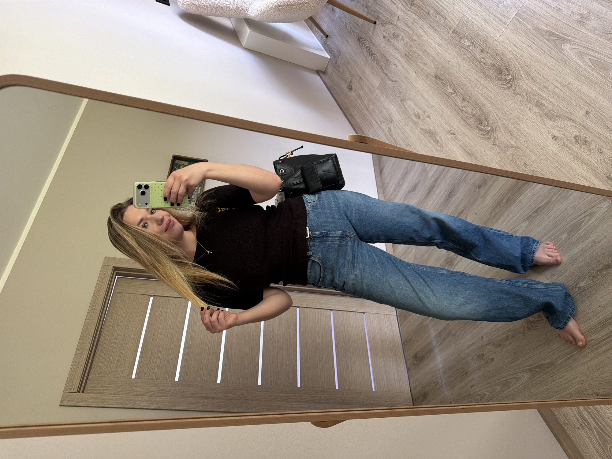 A woman is standing in front of a mirror, taking a selfie with her smartphone. She is wearing a black top and blue jeans. A black purse is slung over her shoulder. She is barefoot. The location appears to be inside a residential building, possibly in Dreiliņi, Latvia, judging from the interior design, including the door. A chair is visible in the background. The floor is wooden.