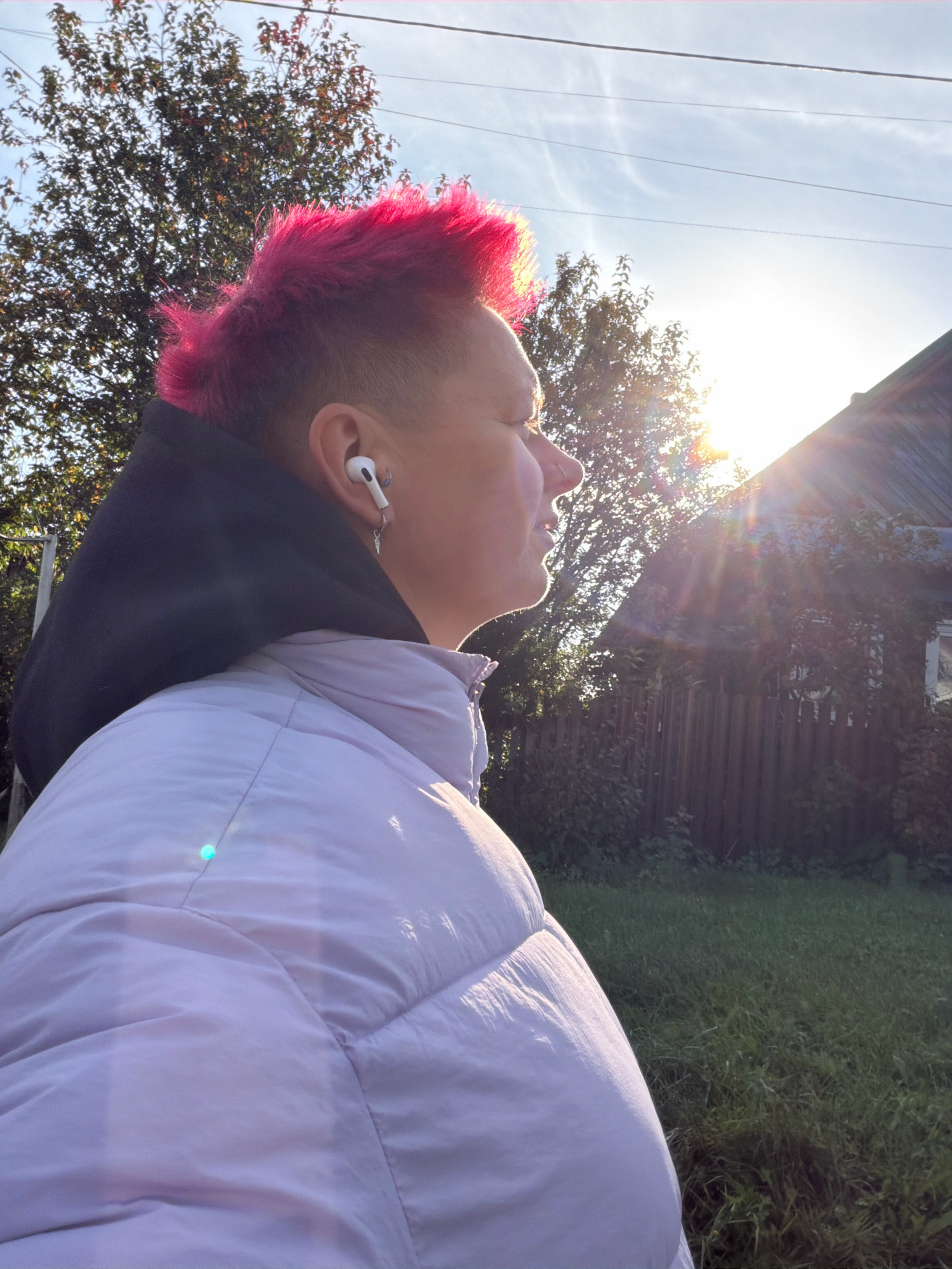 A person with bright pink mohawk hair is seen in profile, looking towards the right of the frame. They are wearing a black hoodie and a light purple puffer jacket. A white wireless earbud is visible in their right ear, along with a silver hoop earring and a dangling charm earring. The background features a tree with red leaves, a wooden fence, a rustic house, and a bright sun with lens flares. The ground is covered in green grass. The image appears to have been taken outdoors on a sunny day, likely in a rural or suburban area in Chaikovsky, Russia.