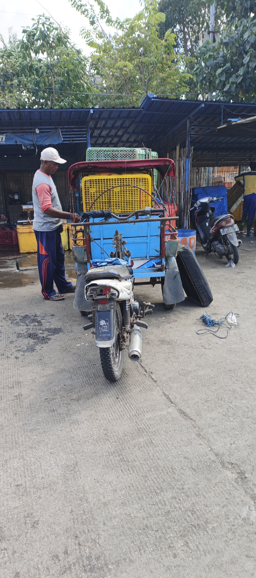 A man wearing a white cap, gray shirt with pink sleeves, and blue pants with red stripes stands by a blue and red tricycle. The tricycle has a yellow basket on top and a white motorcycle attached to the front. The man is holding onto the tricycle's handlebars, and a spare tire is lying on the ground next to it. In the background, a covered market structure is visible, with green trees and a blue sky overhead. A red motorcycle is parked behind the tricycle. Another person in a yellow shirt and blue pants is seen in the far background. The scene appears to be taking place in a market area in Puger, Indonesia.