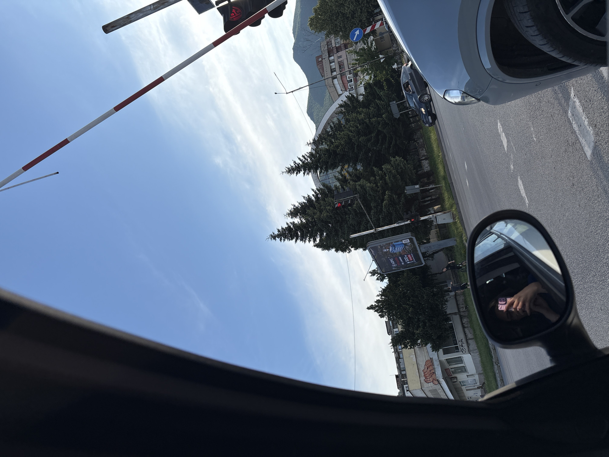 The image captures a view from inside a vehicle in Vratsa, Bulgaria, on a clear day with a bright blue sky and wispy clouds. The foreground is dominated by the interior of the car, including a side mirror reflecting a person holding a pink smartphone.
Outside the car, a street scene unfolds. A red and white striped railway crossing barrier is prominent in the upper right quadrant, tilted upwards. Traffic lights with red lights illuminated are visible on poles. Several tall, dark green coniferous trees line the street. In the background, buildings of varying architectural styles are present, including a modern, curved structure with arched openings. Mountains form the backdrop, suggesting the city's location.
Several vehicles are on the road, including a silver car in the right foreground and a dark sedan in the middle distance. Pedestrians and cyclists are also present, with one person on a bicycle and another walking near a building. A billboard with text and images is positioned to the right of the street. The road itself is asphalt with white lane markings. The overall impression is a snapshot of daily urban life in Vratsa, likely at a railway crossing.
