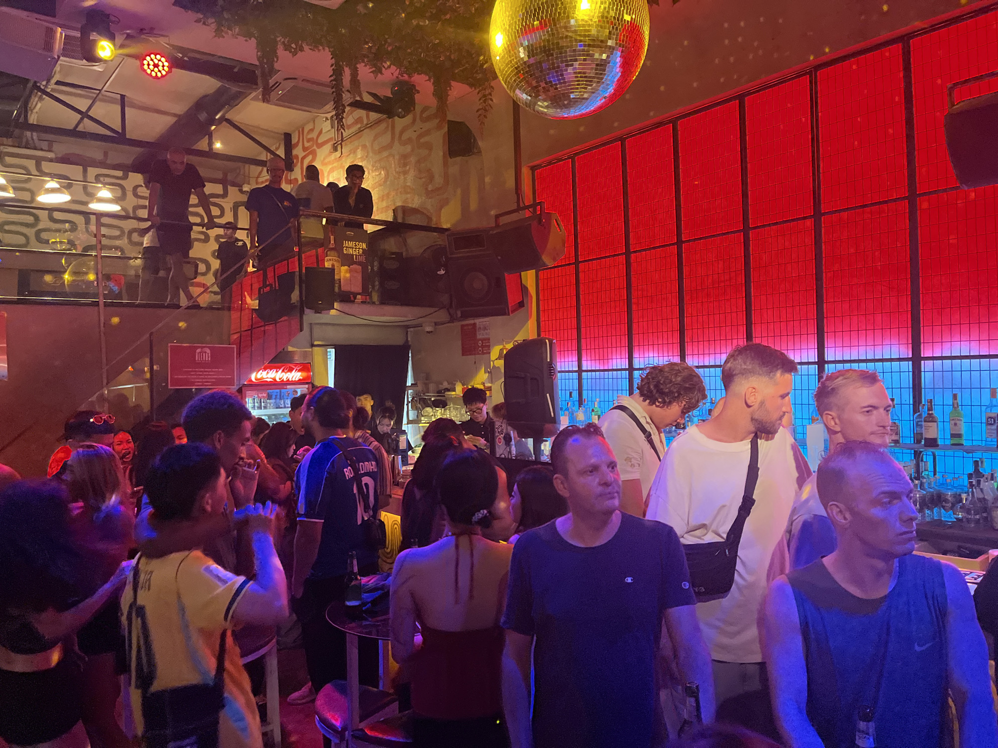 This image depicts a crowded, multi-level bar or nightclub located in Đà Nẵng, Vietnam, bustling with patrons.
The scene is illuminated by vibrant, colored lighting, primarily red and blue, complemented by spotlights emitting yellow and red light, and reflections from a large, gold-toned disco ball suspended from the ceiling. The establishment features an upper mezzanine level with a metal railing and exposed industrial-style beams. On this upper level, a DJ booth is visible on the right, equipped with audio equipment including speakers and a mixer. A male DJ with dark hair stands behind the booth. Beside him, liquor bottles are arranged, including a prominent "JAMESON GINGER LIME" sign. Several individuals, primarily men in casual attire, are present on the mezzanine, with some leaning on the railing overlooking the lower level. On the far left of the upper level, a man in a white t-shirt is partially visible, appearing to be in motion.
The lower level is densely populated with a diverse group of people socializing, standing, and potentially dancing. A long bar counter runs along the right side, stocked with various bottles, some illuminated with blue light from below. Behind the bar, a Coca-Cola refrigerator is visible, along with bartenders serving drinks. In the foreground and midground of the lower level, individuals are engaged in conversation or drinking. Prominent figures include a man in a dark blue Champion t-shirt looking left, a man in a dark blue Nike tank top looking right with a beer bottle in hand, a man in a white t-shirt with a black sling bag, and a man wearing a blue soccer jersey with "RONALDINHO 10" on the back. A woman in a dark red halter top is seated at a small table with a drink, facing away from the viewer. Another man in a yellow soccer jersey is partially visible, also facing away. The red-gridded wall on the right side of the lower level features a strip of blue lighting at its base. Foliage hangs from the ceiling in the upper left section of the venue.
