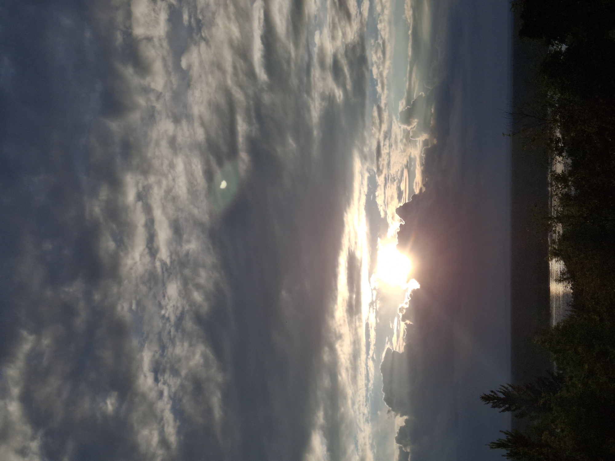 This image depicts a dramatic sky with the sun setting behind clouds. The sun's rays are breaking through, creating a luminous effect that highlights the texture and movement of the clouds. The sky occupies the majority of the frame, with a band of darker clouds on the right side, contrasted by lighter, more wispy clouds stretching across the left.
Below the sky, a body of water is visible, reflecting the light of the setting sun. The water is calm, with a subtle shimmer on its surface. On the far right, the silhouette of trees and foliage frames the scene, suggesting a natural landscape. The overall impression is one of a serene yet powerful natural spectacle.
The time of day is clearly sunset, indicated by the low angle of the sun and the warm light illuminating the clouds and water. The weather appears to be partly cloudy, with dramatic formations creating visual interest. While no specific location cues are present in the image itself, given the context of Chaikovsky, Russia, it could be a view over the Kama River or a similar body of water. The scene evokes feelings of awe and tranquility. No text is visible in the image.