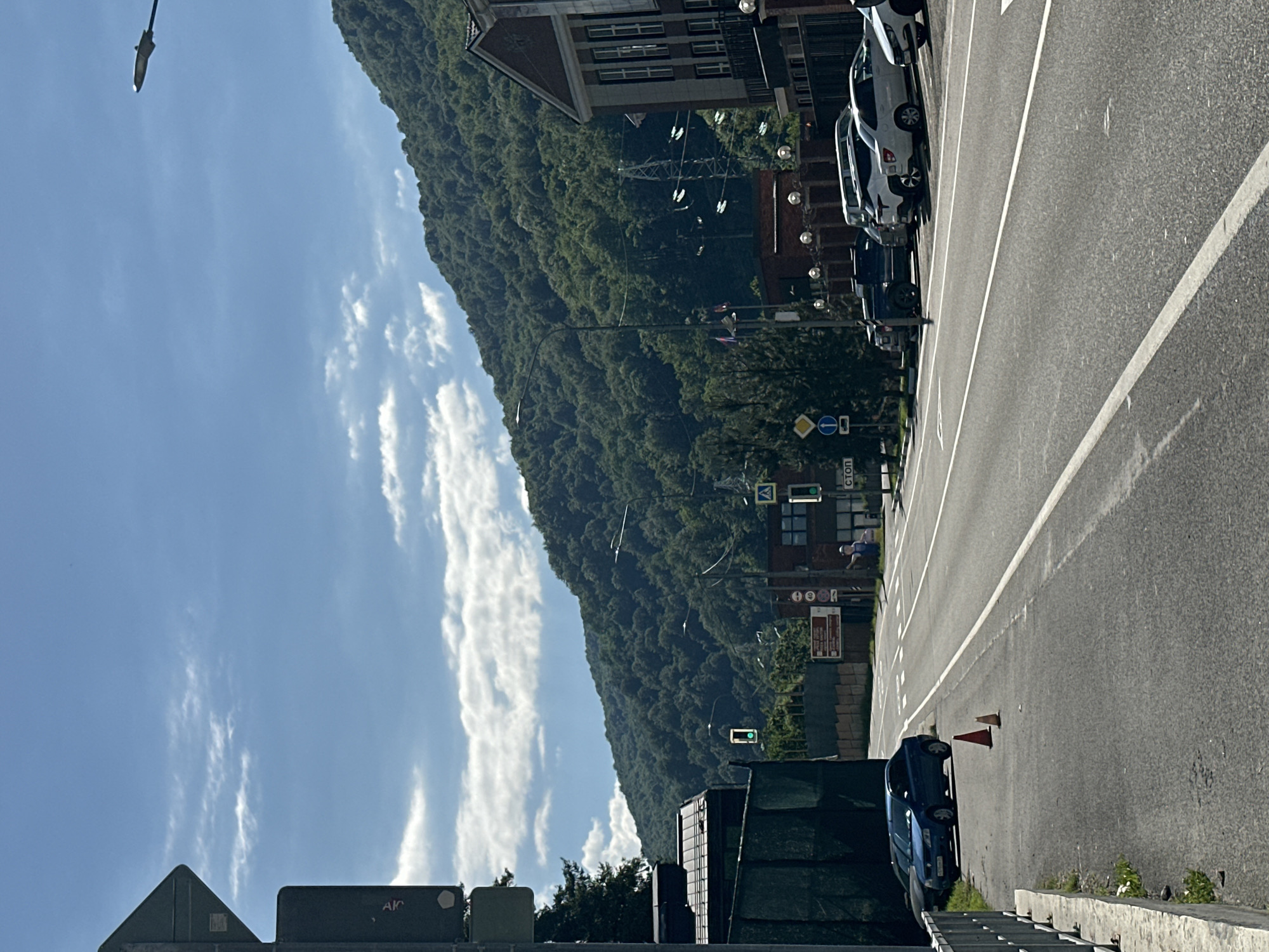 The image depicts a vibrant daytime street scene set against a backdrop of a large, lush green mountain, characteristic of Krasnaya Polyana, Russia. A wide asphalt road, marked with clear white lines, stretches from the foreground into the distance on the right side of the frame.
On the right side of the road, several cars are parked, including multiple white vehicles and a dark blue sedan closer to the foreground. Two orange traffic cones are placed near the blue car. Along the roadside, there are multi-story buildings featuring brick and concrete facades, some with visible windows.
Traffic infrastructure is prominent, with streetlights, power lines, and various road signs visible. A pedestrian crossing is marked on the road, accompanied by traffic lights, one of which shows a green signal. In the mid-ground, a person wearing light-colored clothing can be seen walking on the sidewalk near the crossing.
The weather appears bright and sunny, with a clear blue sky scattered with wispy white clouds. The time of day is clearly daytime, likely morning or early afternoon, given the bright illumination and visible shadows.
Visible Cyrillic text includes "СТОП" (STOP) on several road signs. The letters "АК" (AK) are also discernible on a dark sign in the lower left foreground. Other signs contain illegible Cyrillic text.