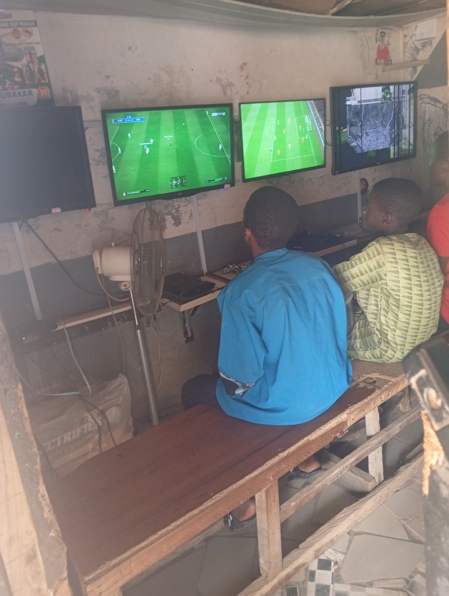 The image depicts an indoor setting, possibly a makeshift gaming center, in Jos, Nigeria. Three screens are visible, two displaying soccer video games and one showing a map. Two young boys are seated in front of the screens, engaged in playing the games. A floor fan is positioned between the boys, suggesting the environment might be warm. The walls are a textured, off-white, and the ceiling appears to be constructed from corrugated metal. Posters with text and images are affixed to the wall, one of which seems to be a political poster. Various electronic devices and cables are present, indicating the operation of the gaming setup. The overall scene suggests a casual, recreational space, likely a local business catering to video game enthusiasts.