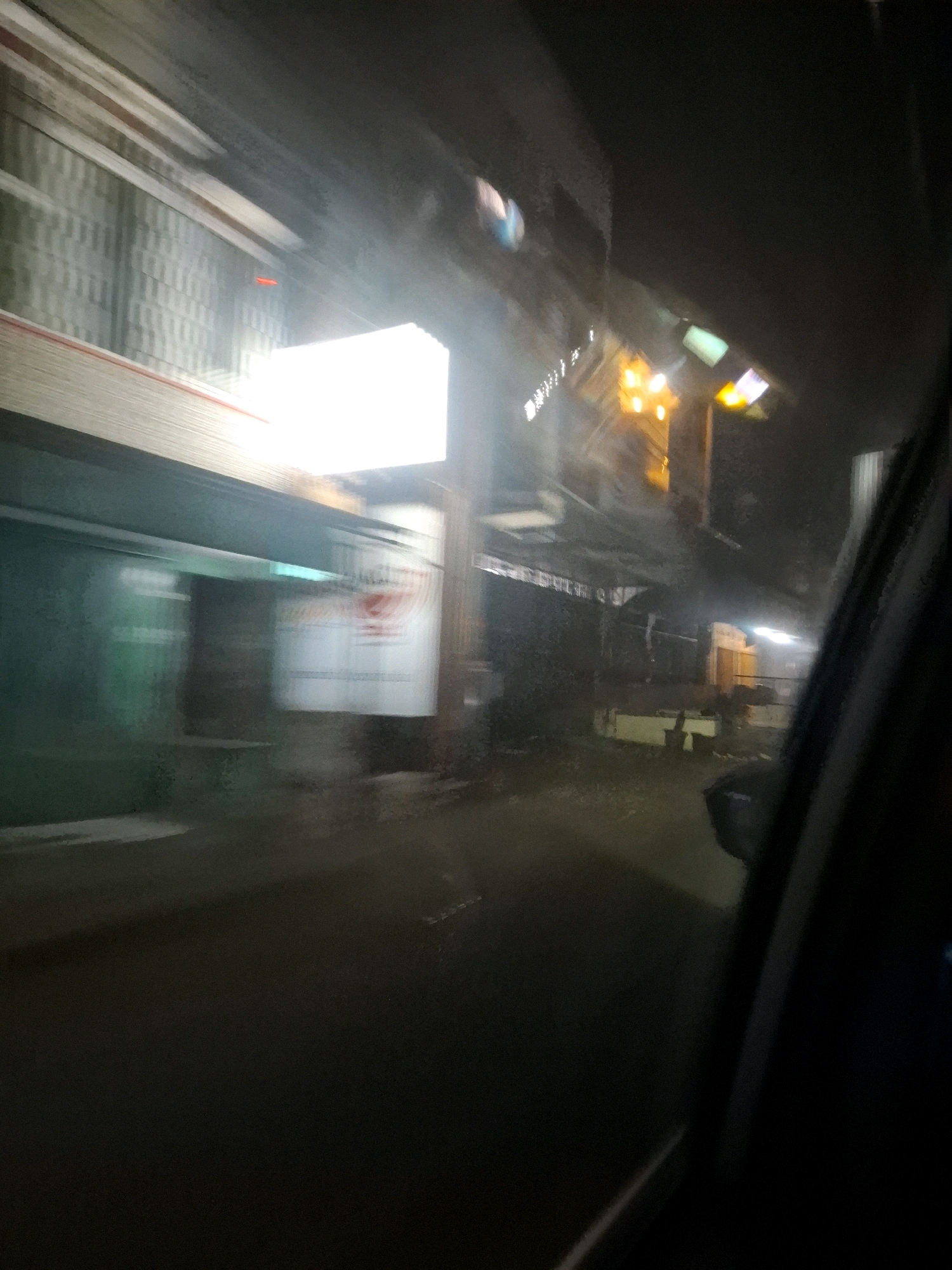 This is a blurred, nighttime photograph taken from inside a vehicle, likely in motion, offering a view of a street scene in Nagrak, Indonesia. The foreground is dominated by the dark interior of the vehicle, with the right side of the frame showing a dark car door or pillar. The environment outside appears to be an urban or semi-urban area with some commercial buildings.
On the left side of the image, there is a long, light-colored structure with horizontal lines, possibly a building facade or scaffolding. Below this, a green-colored structure with blurry horizontal lines is visible, suggesting a shop front or a wall. A large, illuminated rectangular sign with red text on a white background is present in the middle of the frame, heavily blurred due to motion. Further to the right, a brightly lit orange object, possibly a street lamp or neon sign, is visible, casting a warm glow. Another illuminated sign or light fixture, appearing greenish or bluish, is seen above and to the left of the orange light.
The ground appears to be a dark, wet surface, suggesting rain or damp conditions, with some indistinct shapes and reflections. There are also structures resembling fences or railings in the background. The overall impression is of a transient view of a dimly lit street with commercial activity at night.