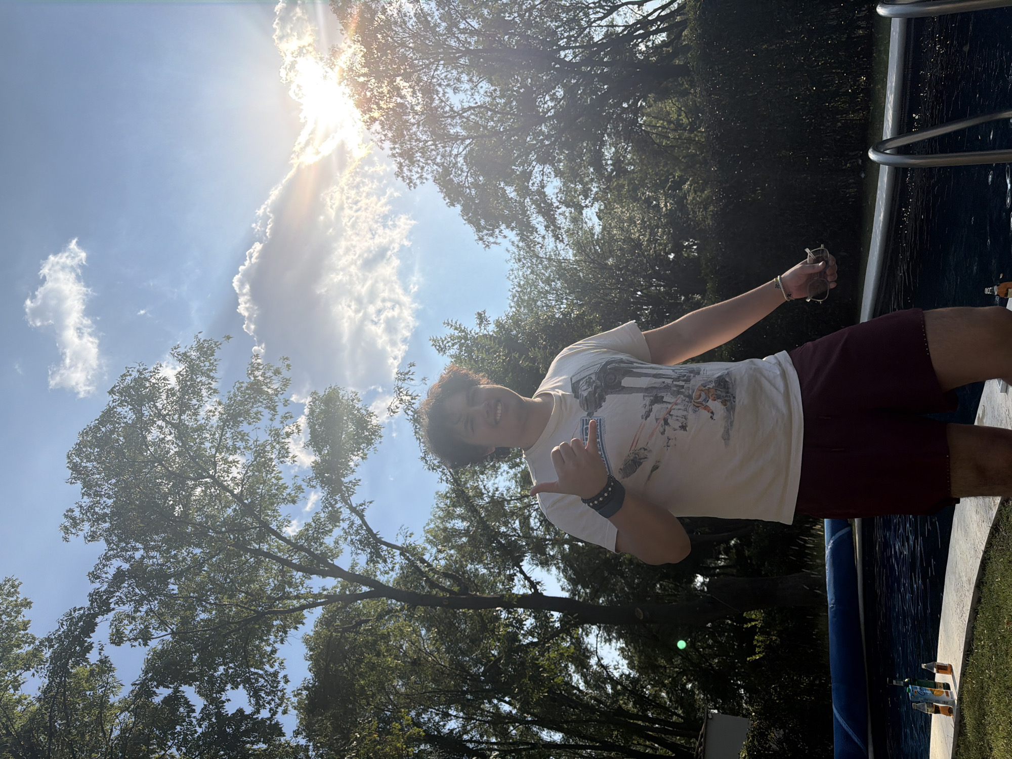 A young person stands outdoors, near what appears to be the edge of a pool, based on the presence of water, pool rails, and bottles near the edge. The individual is wearing a light-colored t-shirt and dark shorts. The person is gesturing with their left hand, possibly making the "shaka" sign, and holding an object in their right hand, possibly sunglasses and a phone. The background shows trees and a bright, slightly cloudy sky with the sun shining. The location is Santa Ana Yancuitlalpan (Chilhuacán), Mexico.